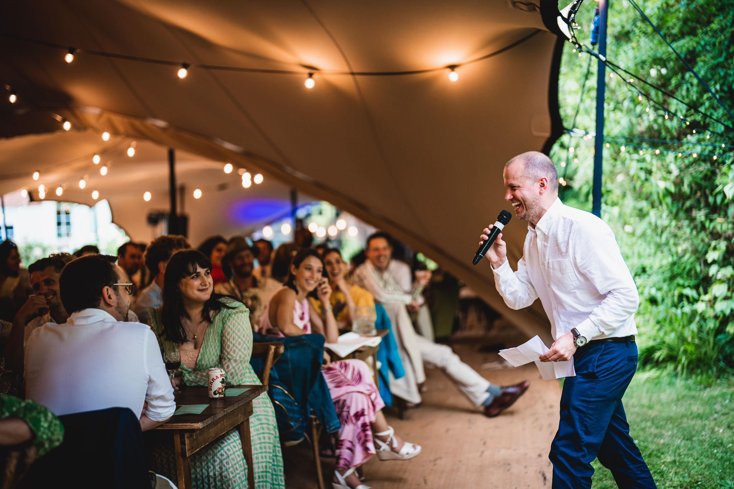 A person in a white shirt gives a speech with a microphone under a tent at an event, with seated guests listening and smiling.