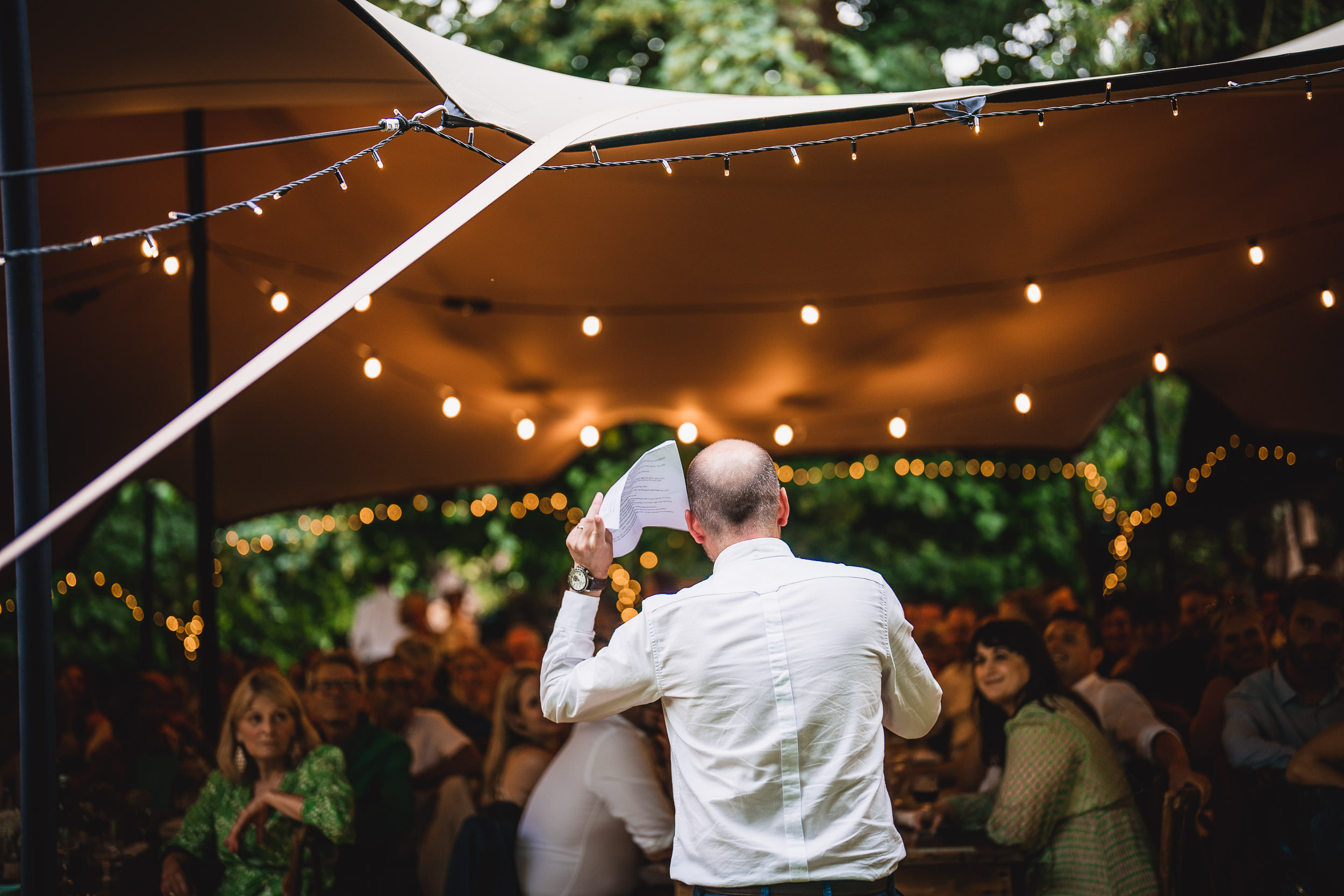 A person holding a paper speaks to an audience seated under a tent with string lights in a lush outdoor setting.