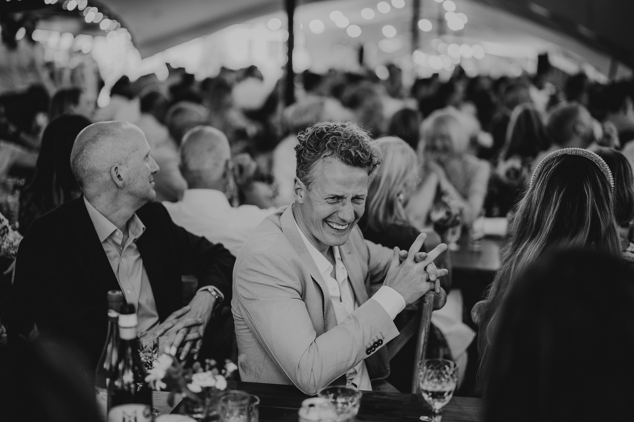 A man in a suit laughs at a crowded indoor event. Tables are set with drinks and flowers. The room is filled with blurred people and string lights in the background.