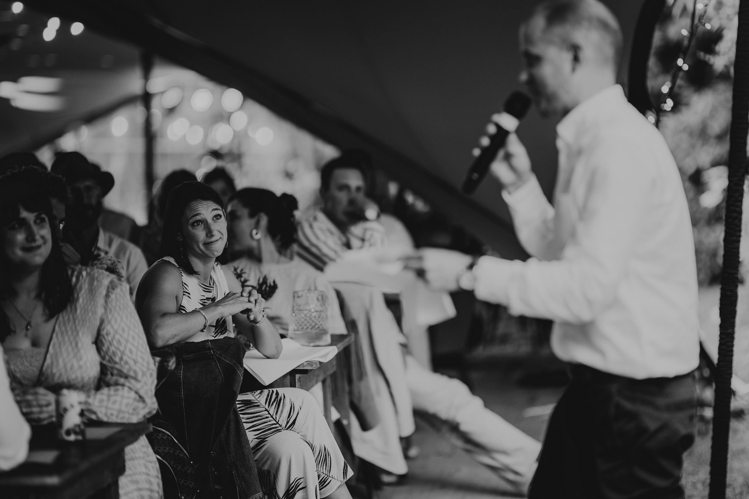 A black and white photo of a seated audience listening to a person speaking into a microphone at an indoor event. One woman looks toward the speaker with an attentive expression.