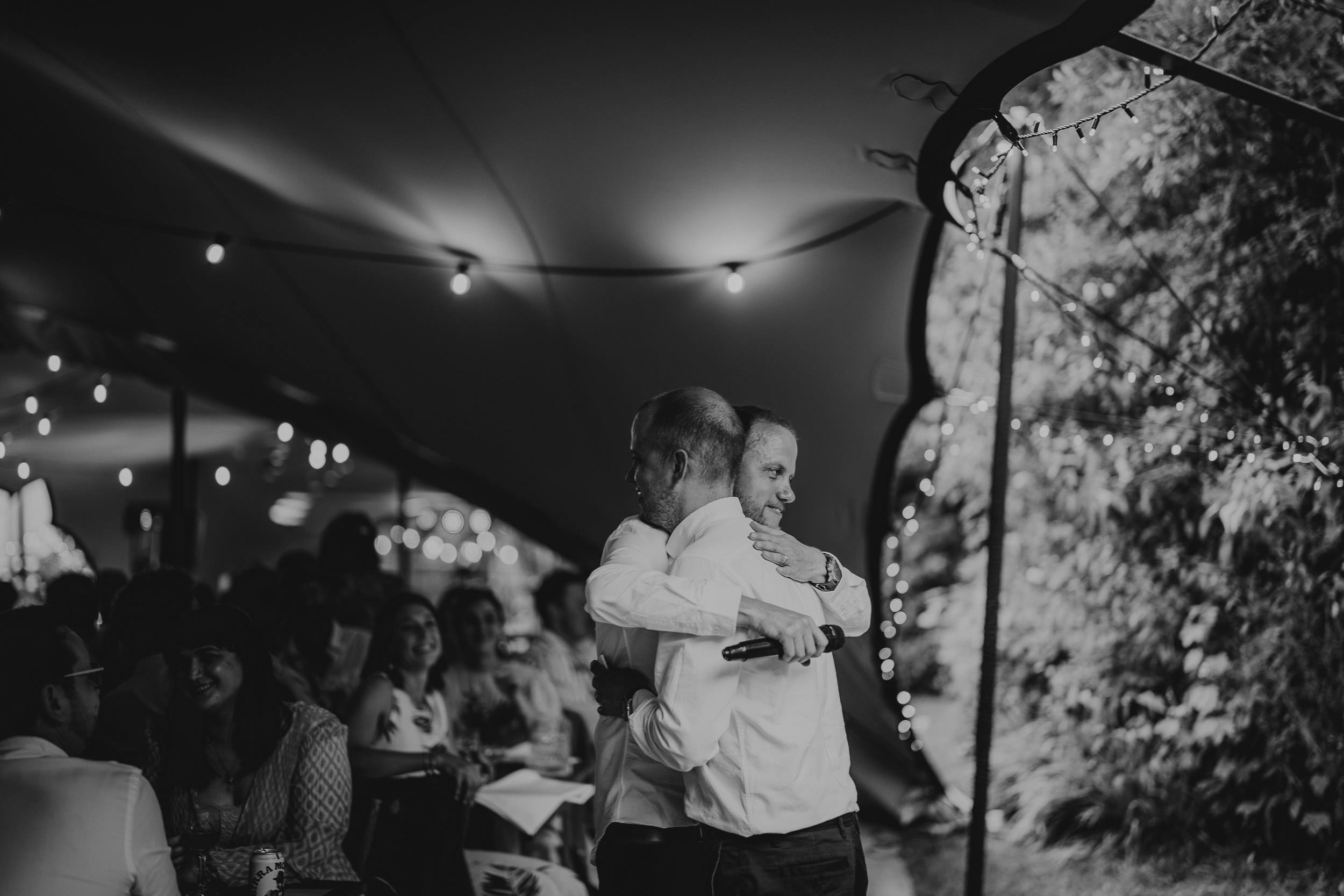 Two people hug under a tent adorned with string lights, surrounded by seated guests at an event.