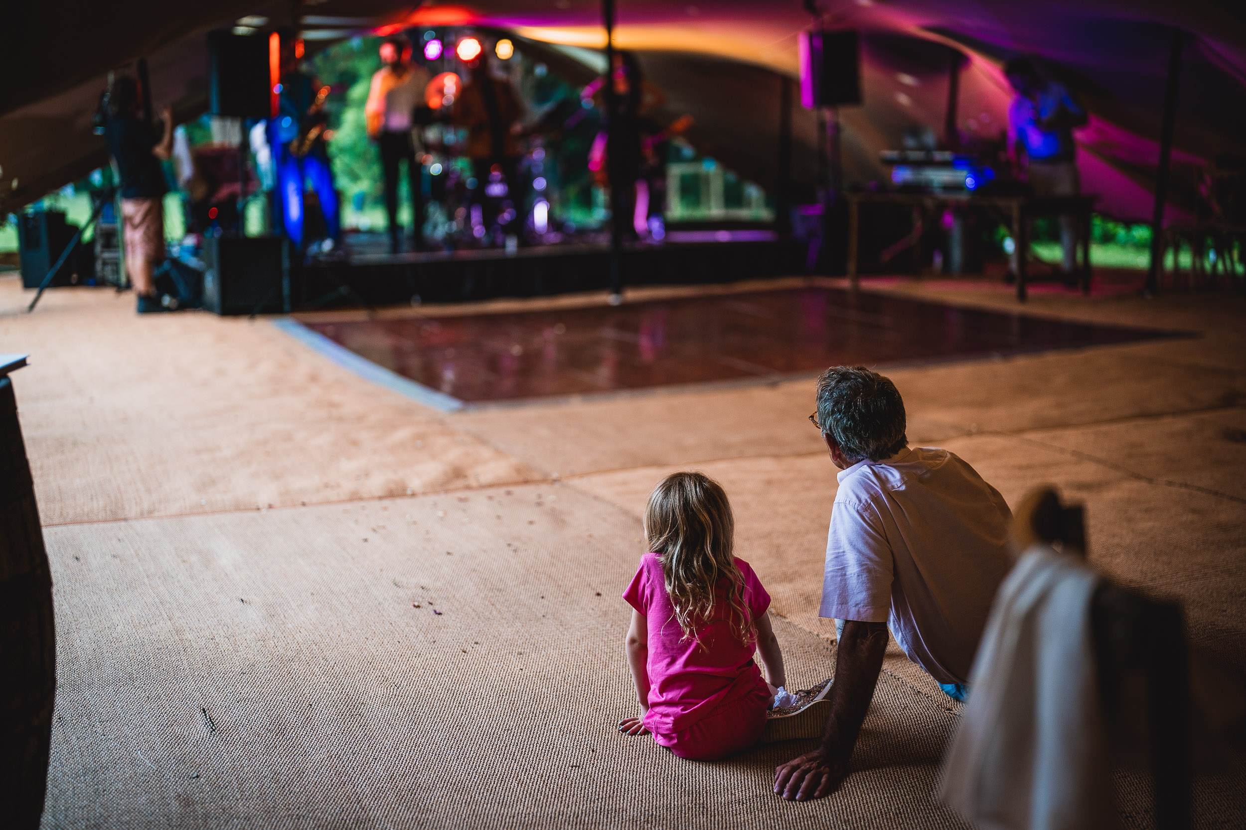 A man and a young girl sit on the ground inside a tent, watching a band perform on stage under colorful lights.