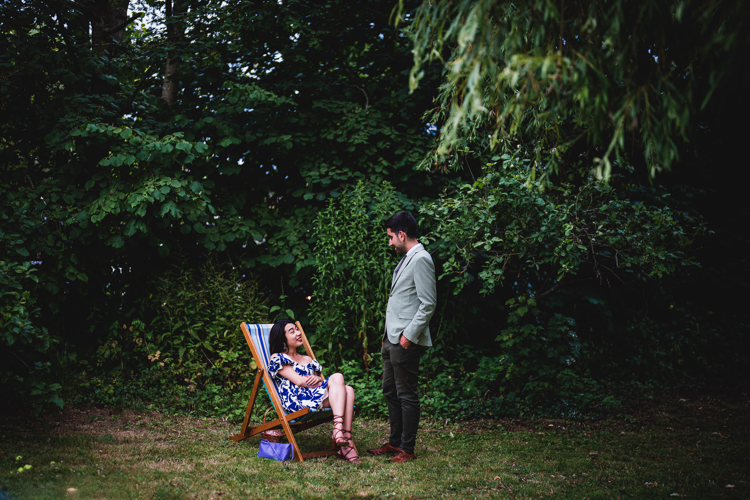 A woman in a patterned dress sits on a deck chair in a garden, looking at a man in a gray blazer and dark pants standing in front of her.