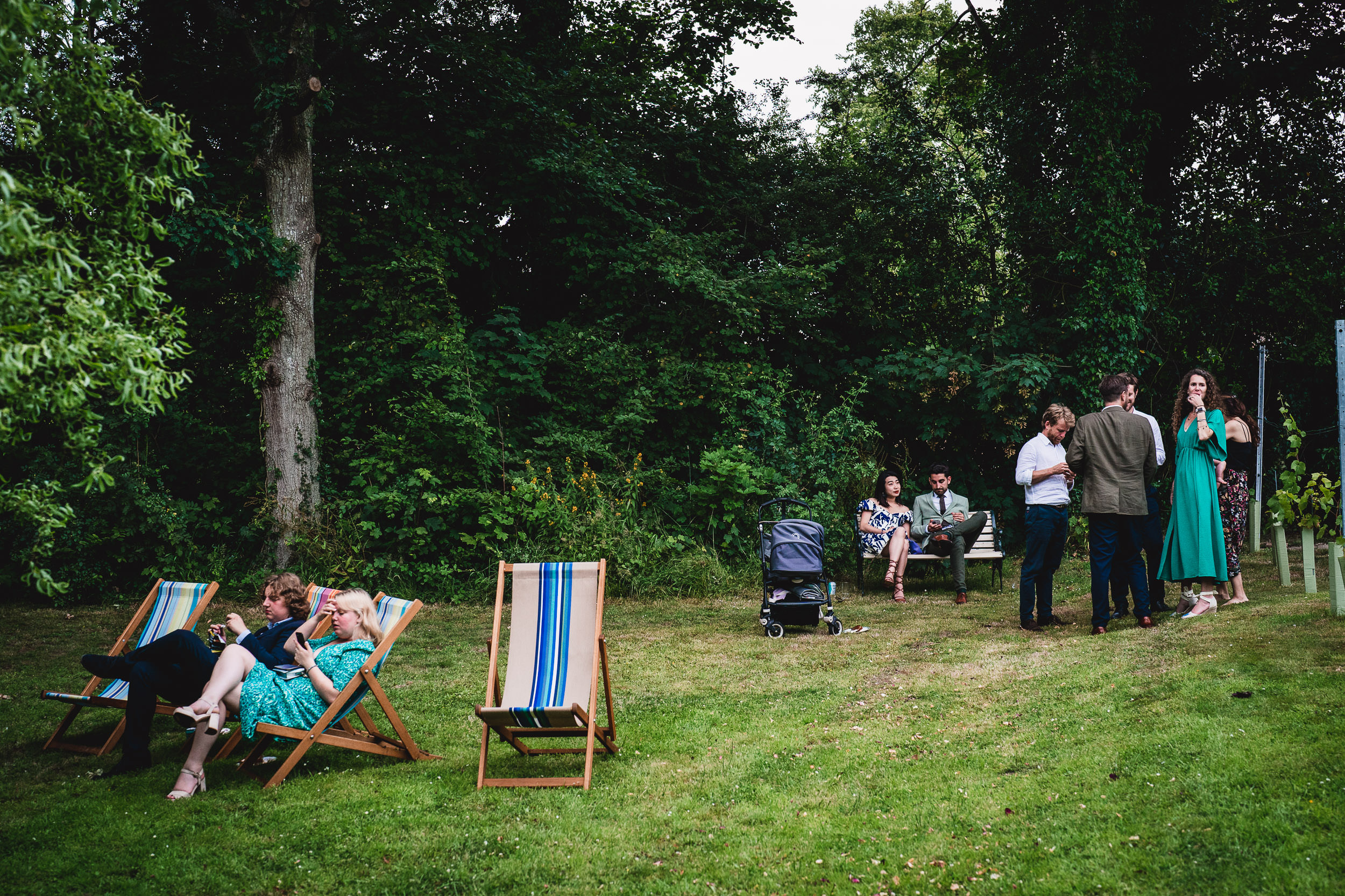 People relaxing in a garden, with some seated on deck chairs and others standing. A stroller and trees are in the background.