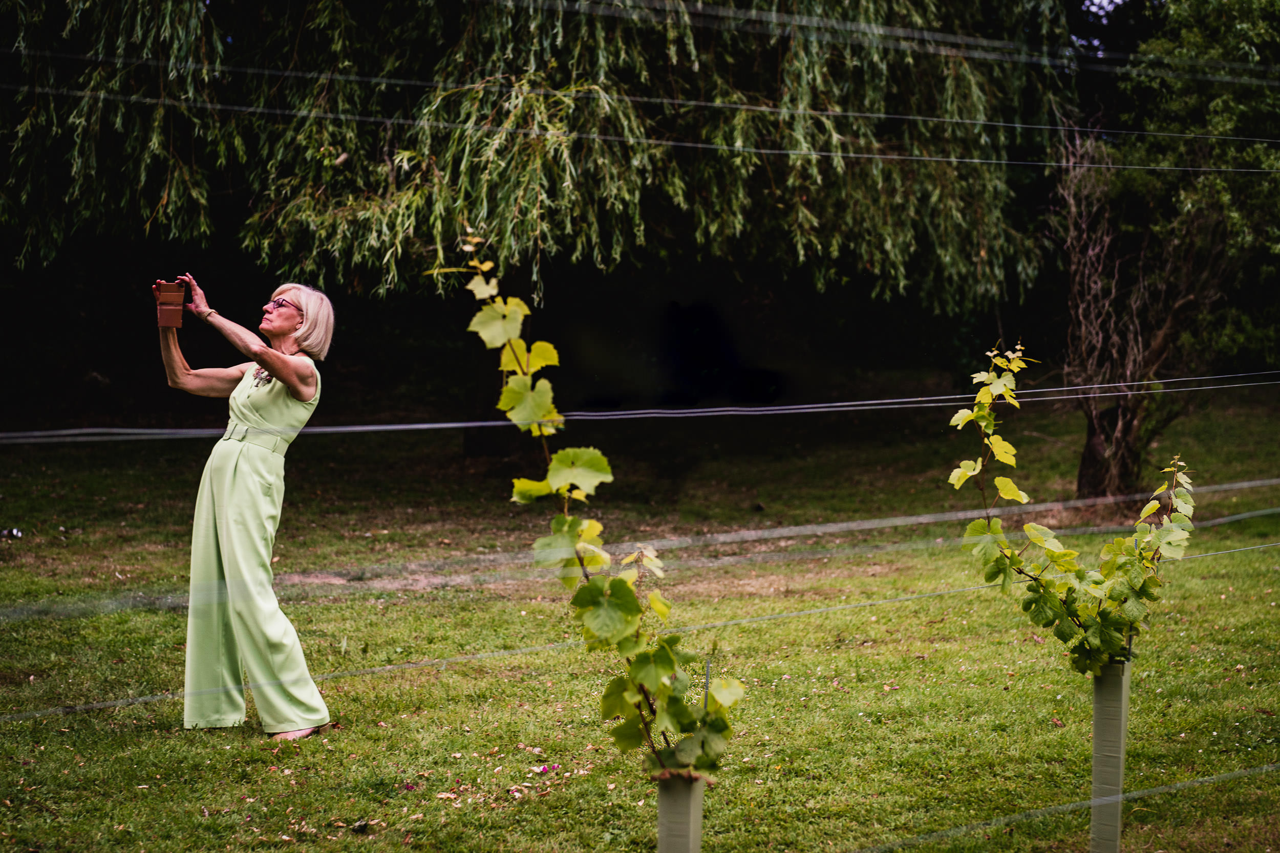 A woman in a light green dress takes a selfie in a garden with small plants and trees in the background.