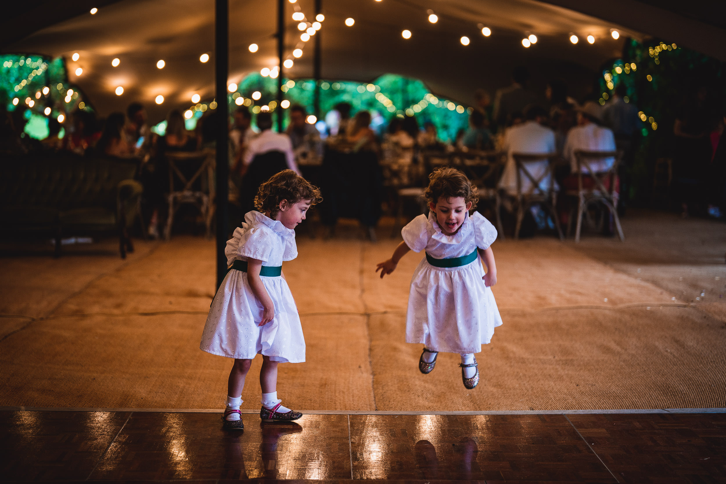 Two young children in white dresses play joyfully under string lights at a festive gathering.