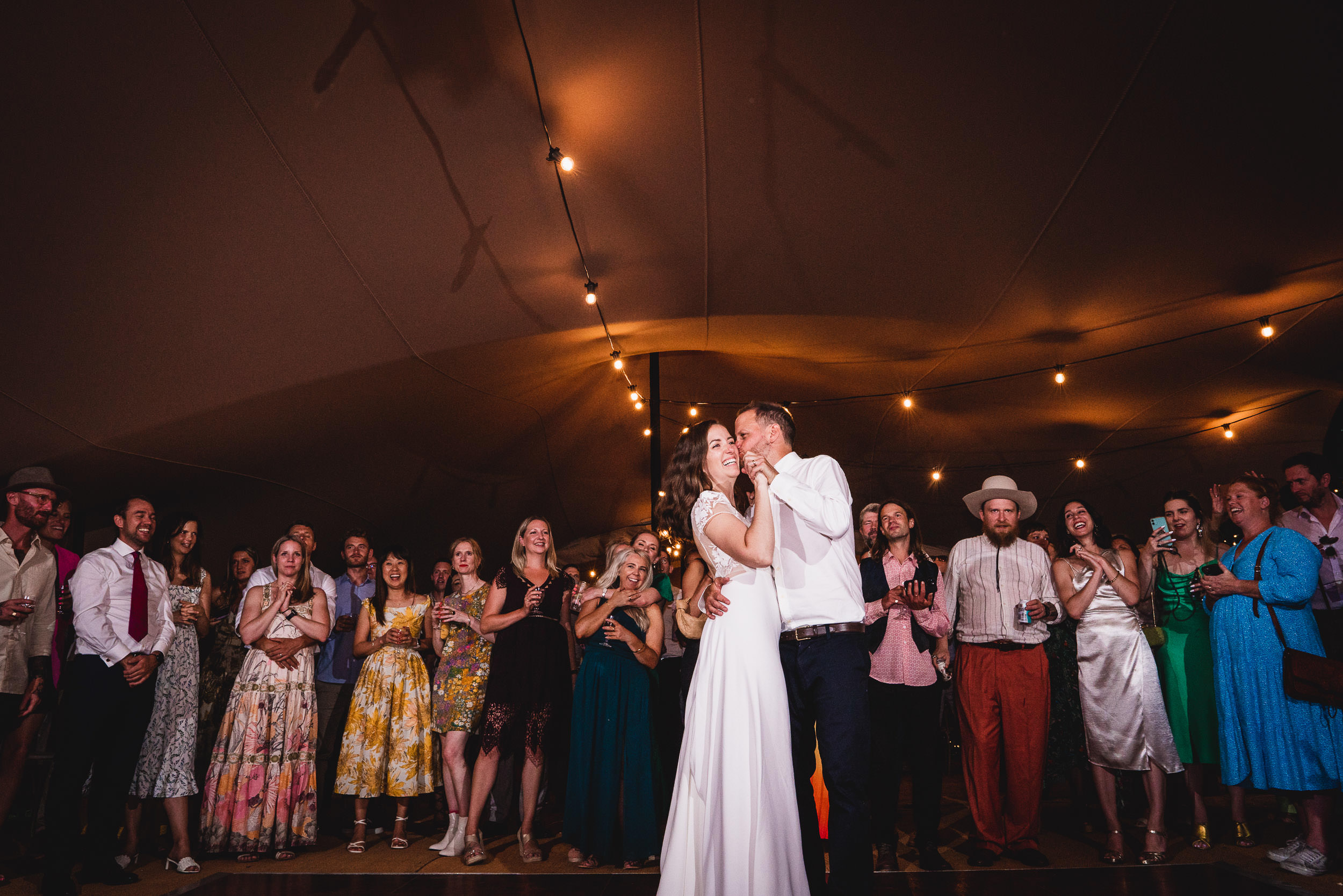 A couple dances under string lights in a tent surrounded by a crowd of people watching and smiling.