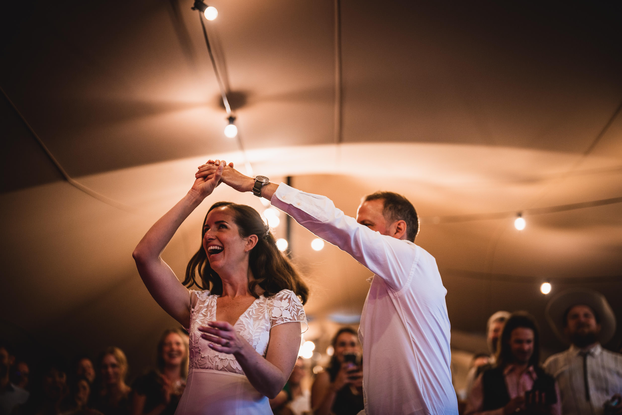 A couple dancing joyfully under string lights at an indoor event, surrounded by smiling guests.