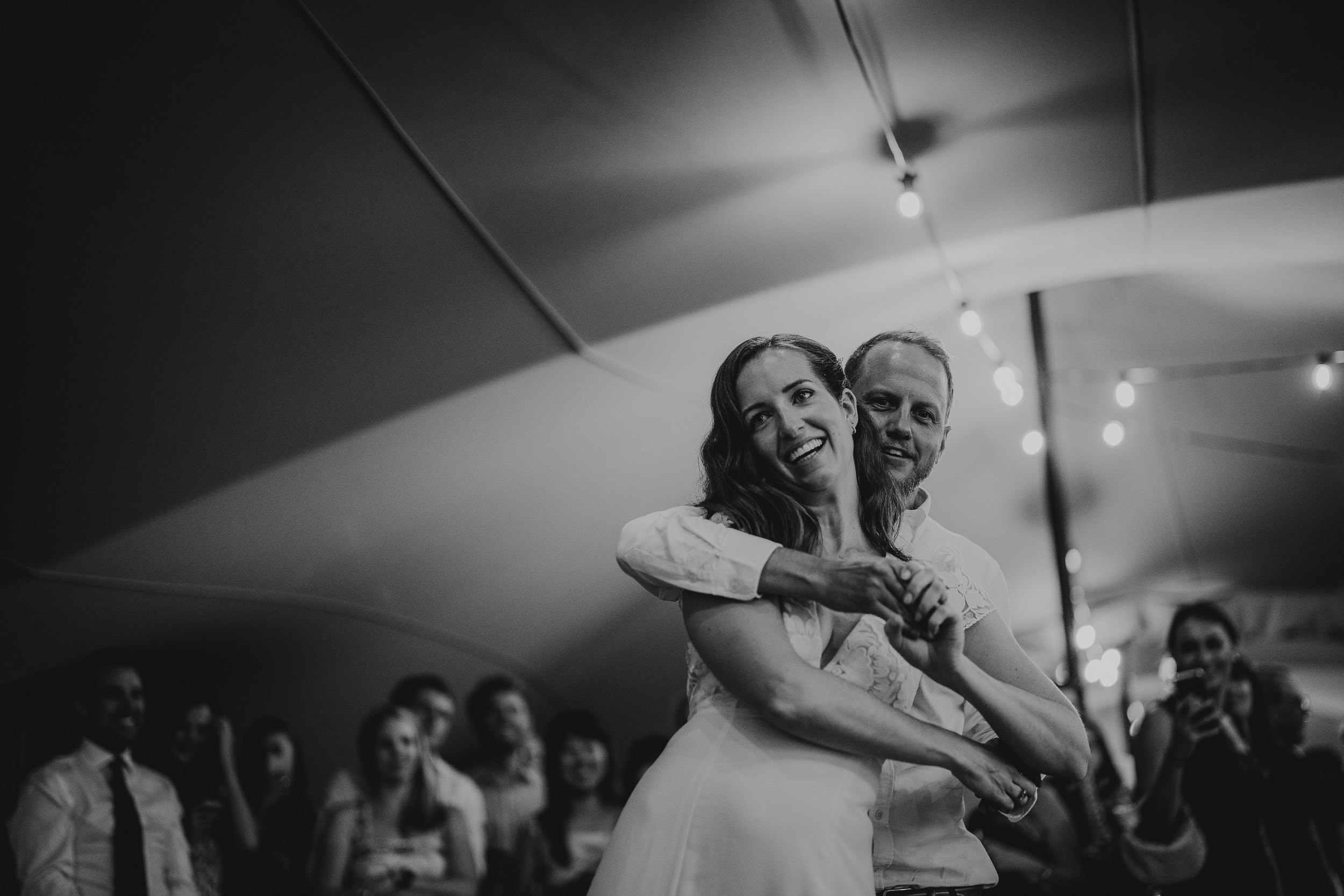 Bride and groom dancing joyfully under string lights at a wedding reception, surrounded by guests in a tent.