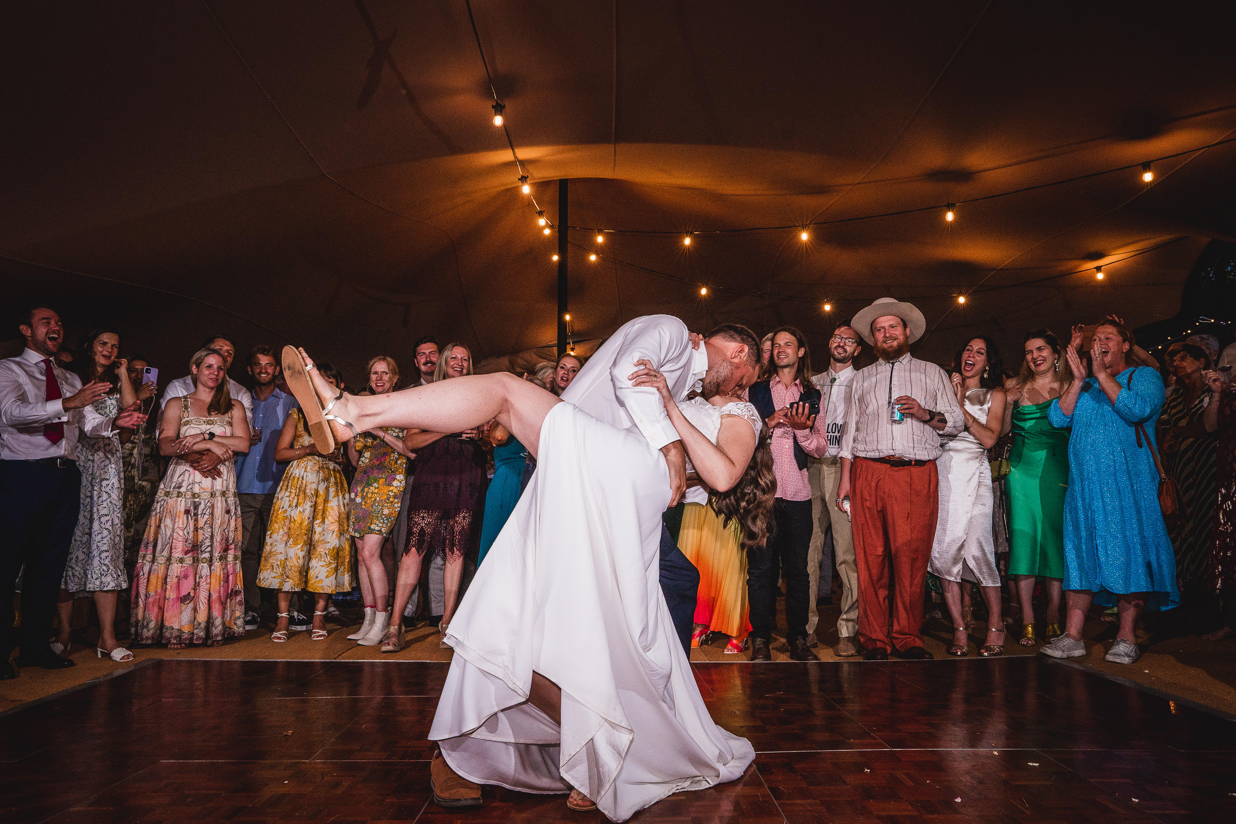 A couple kisses on the dance floor during a wedding reception, surrounded by cheering guests under string lights.