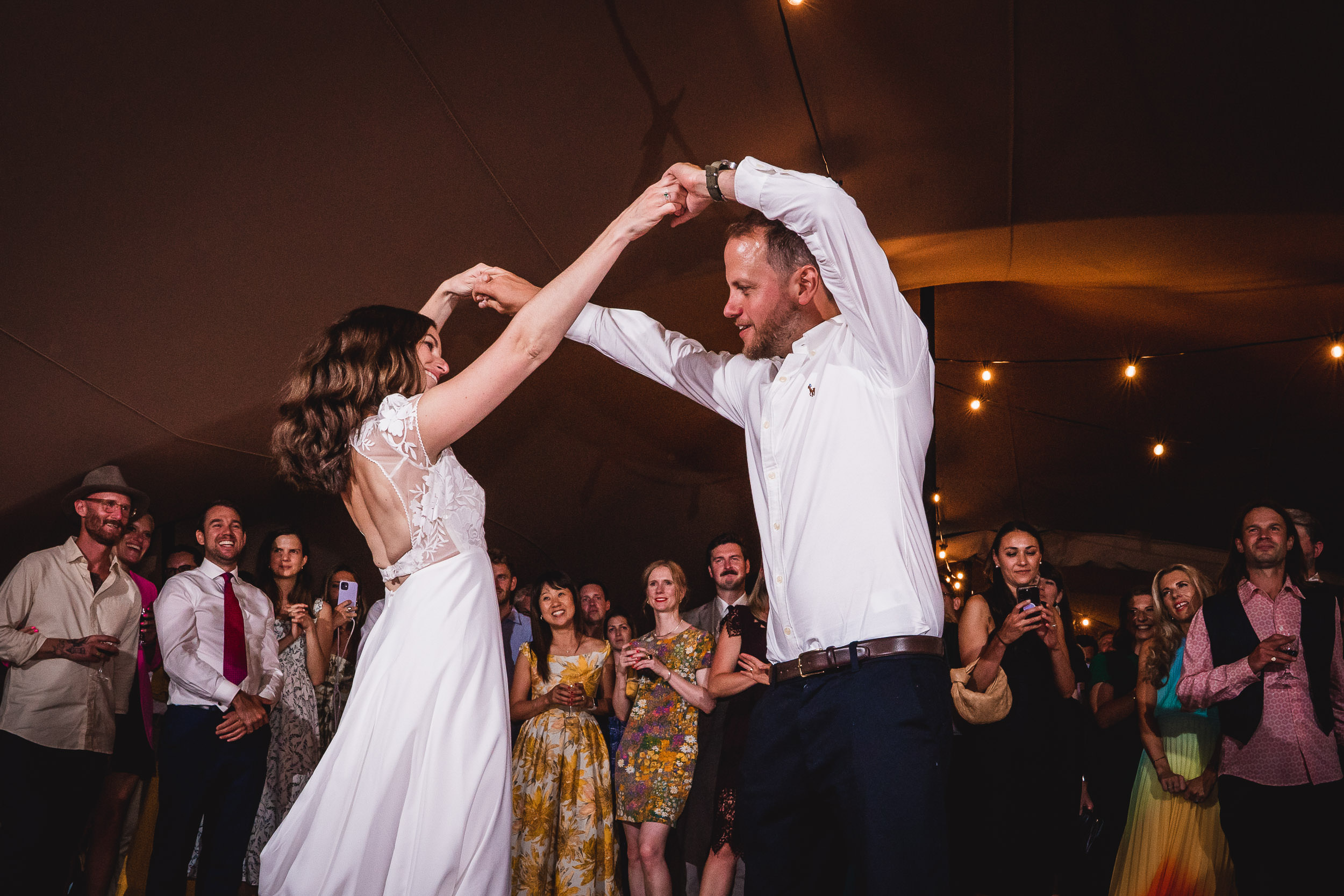 A couple dances under string lights at a wedding reception. A group of guests stands around them, watching and smiling.