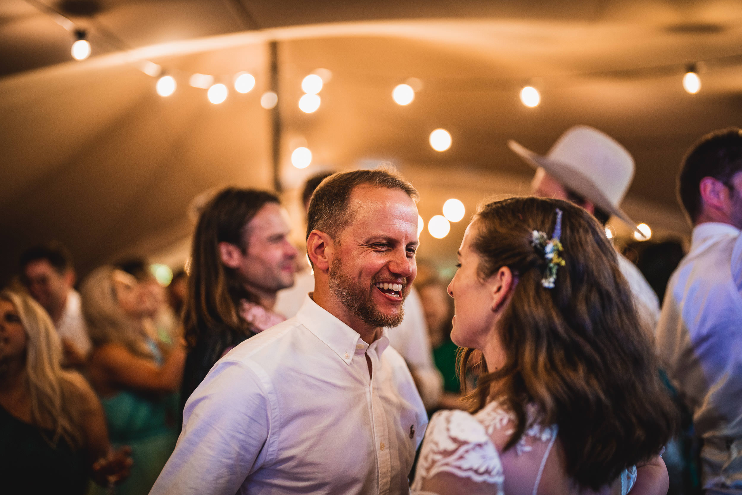 A man and woman smiling at each other in a warmly lit venue, surrounded by people and decorative lights.