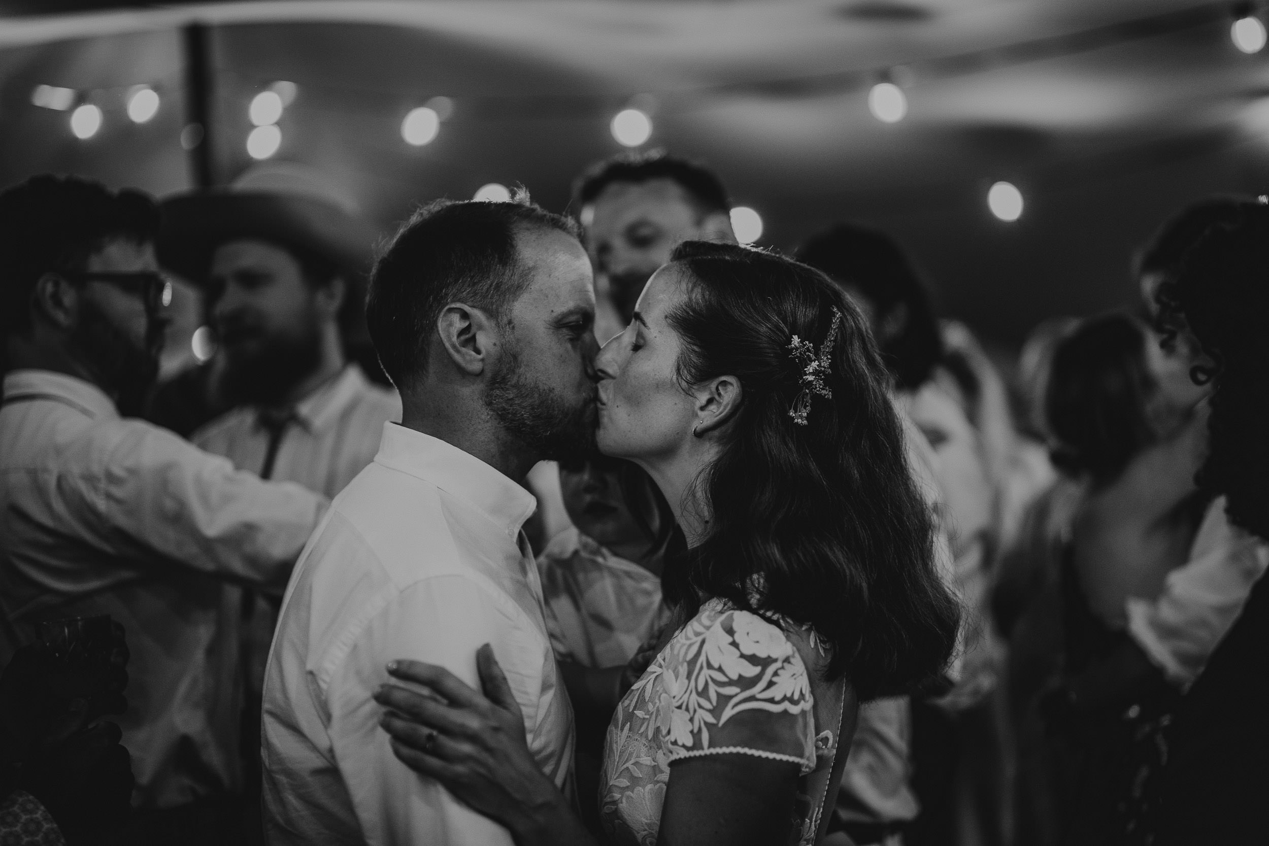 A couple kisses on a dance floor, surrounded by people in a dimly lit setting with string lights above.