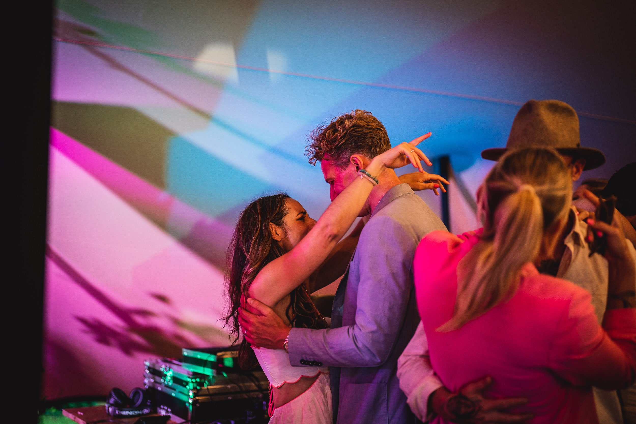 A couple dances closely under colorful lights at a lively party, surrounded by other people.