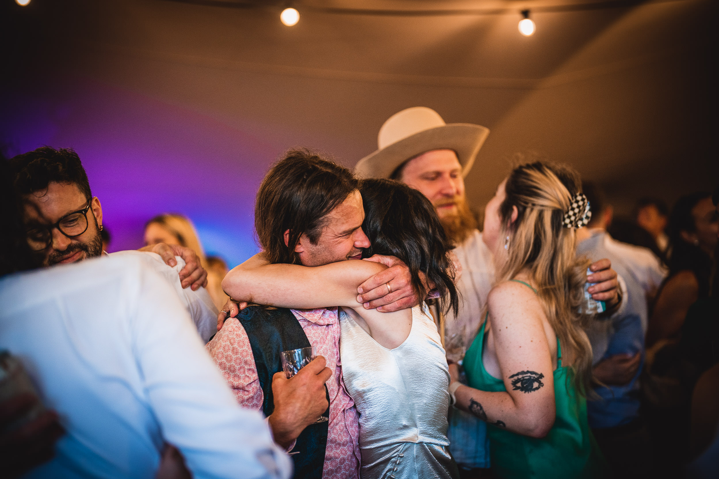 A group of people at a lively party. A couple in the center embraces; others stand nearby, one wearing a cowboy hat. The atmosphere is joyful with colorful lighting.