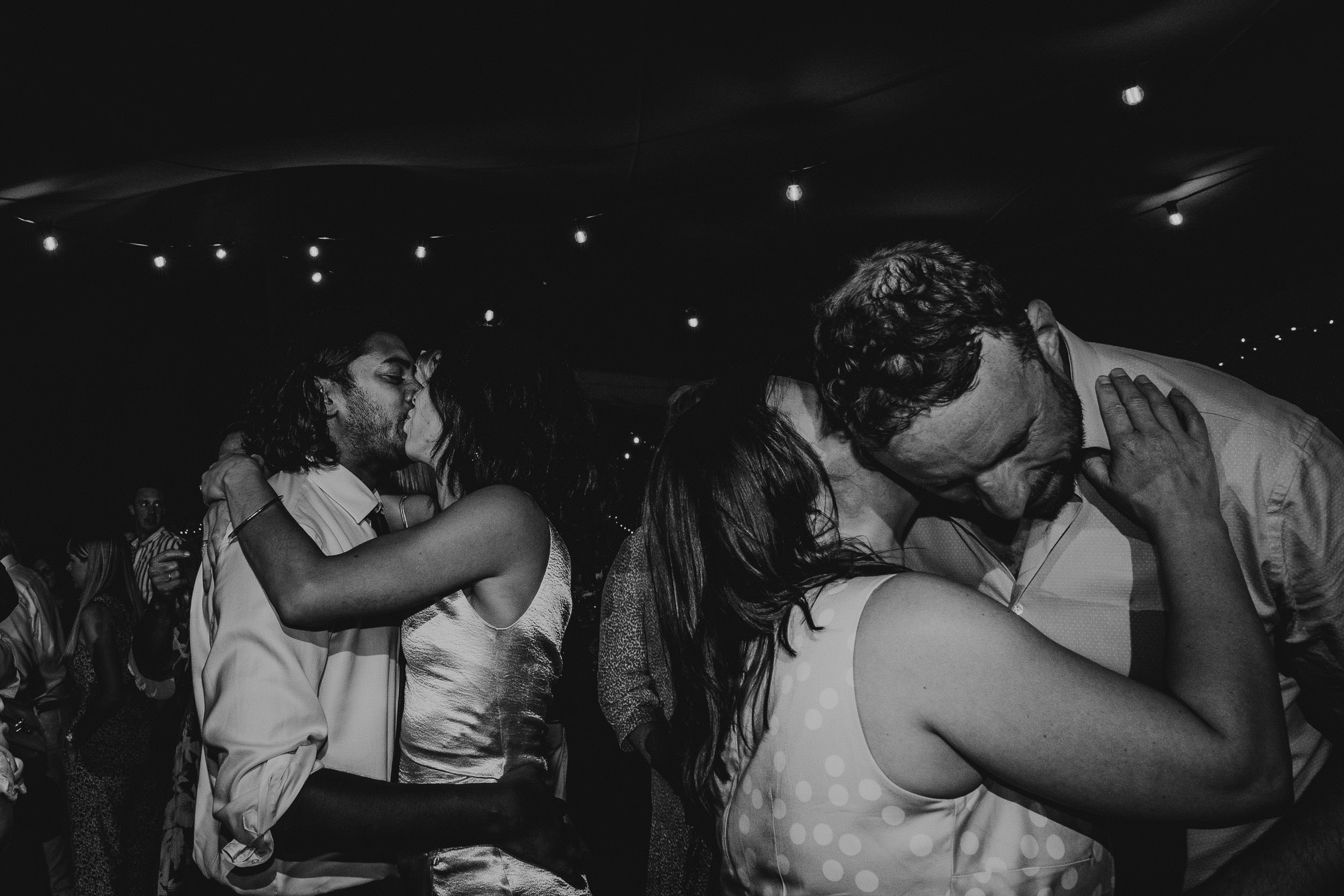 Two couples are kissing and embracing under string lights at a nighttime event.