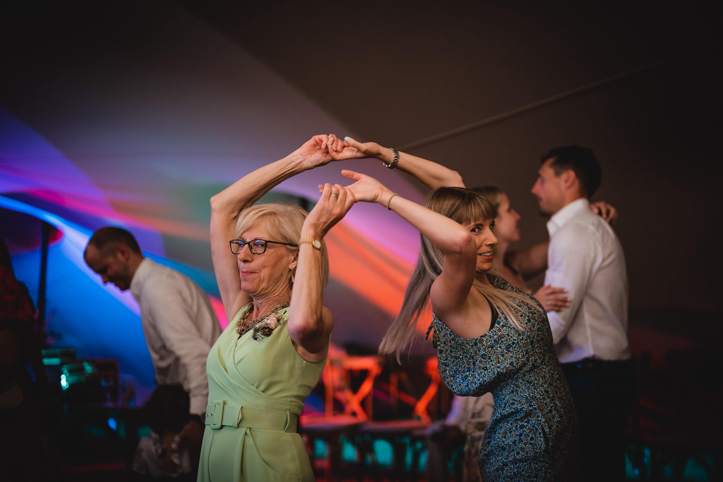 Two women are dancing together, holding hands, in a colorful, softly lit room. People in the background are also dancing.