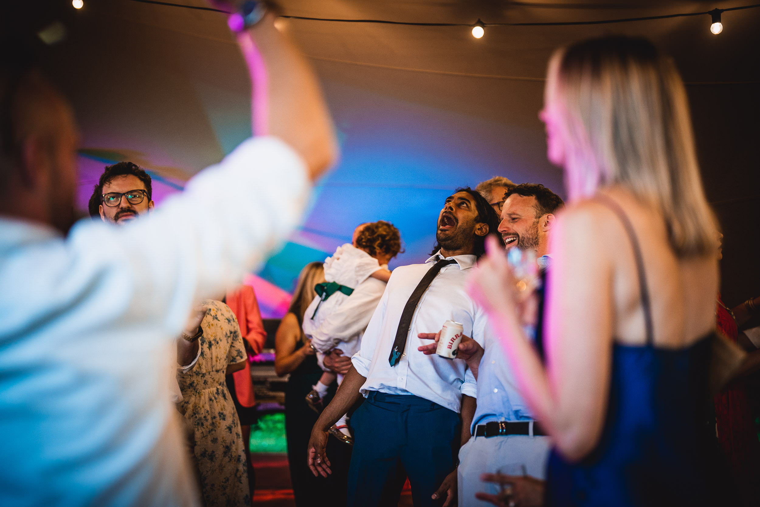 People enjoying a lively celebration under colorful lights, some holding drinks, with joyful expressions and festive attire.