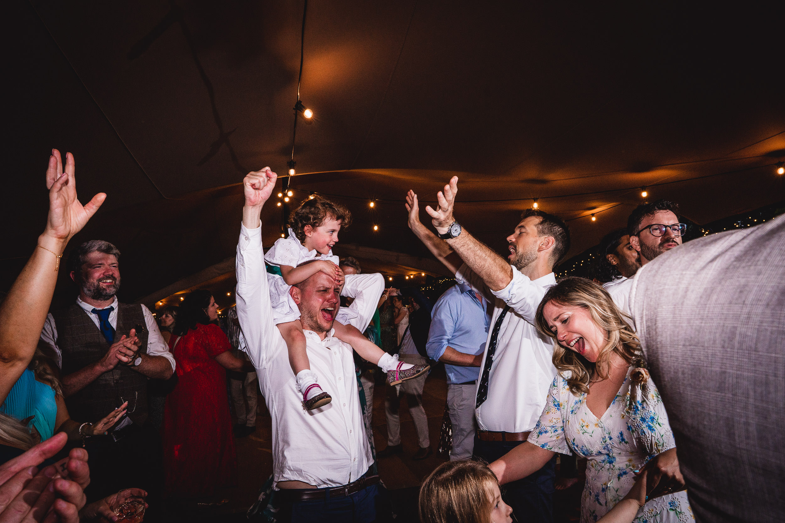 A joyful group dances under string lights; a man holds a child on his shoulders, while others cheer with raised arms.