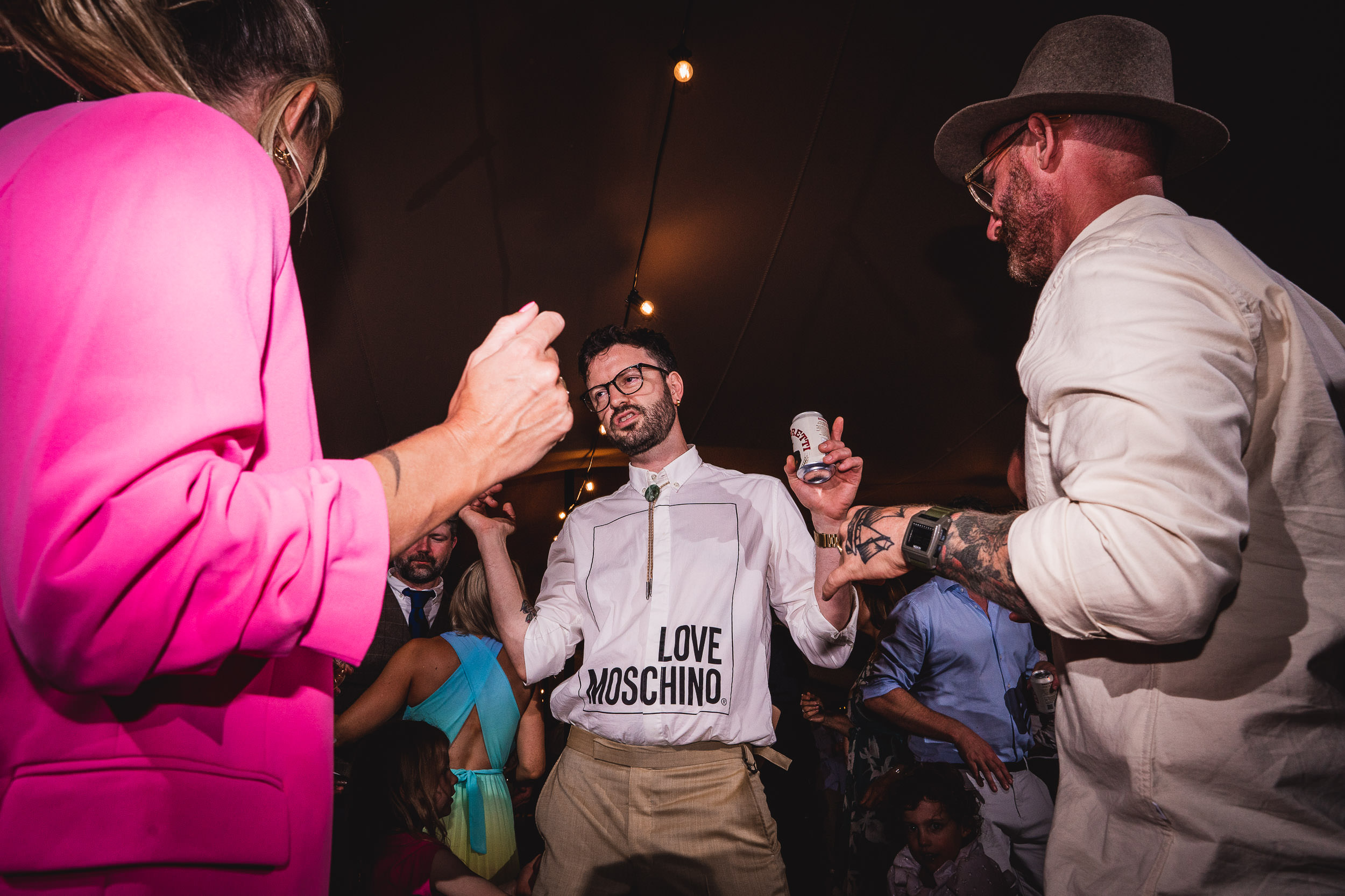 A group of people dancing at a party, one man wearing a "Love Moschino" shirt and glasses, holds a can. Another man in white and a hat stands nearby.