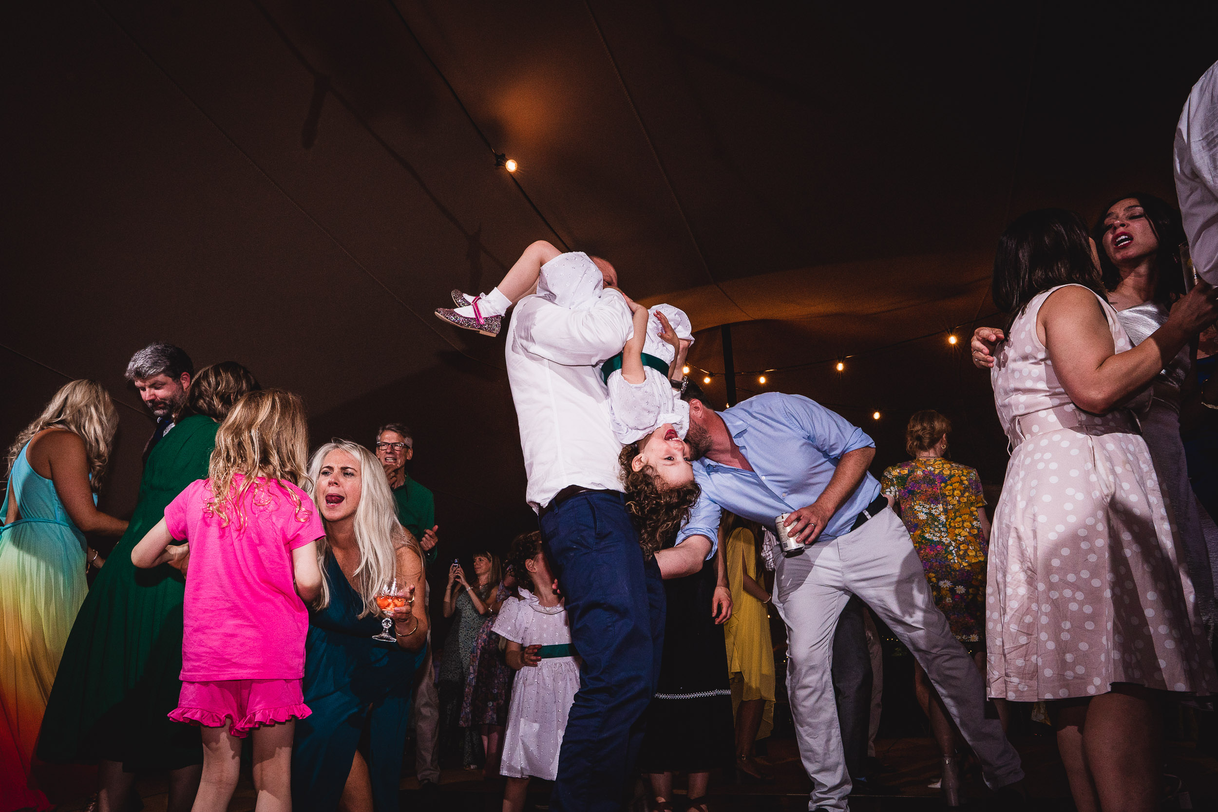 A lively crowd dances at an indoor event; one person playfully holds a child upside down.