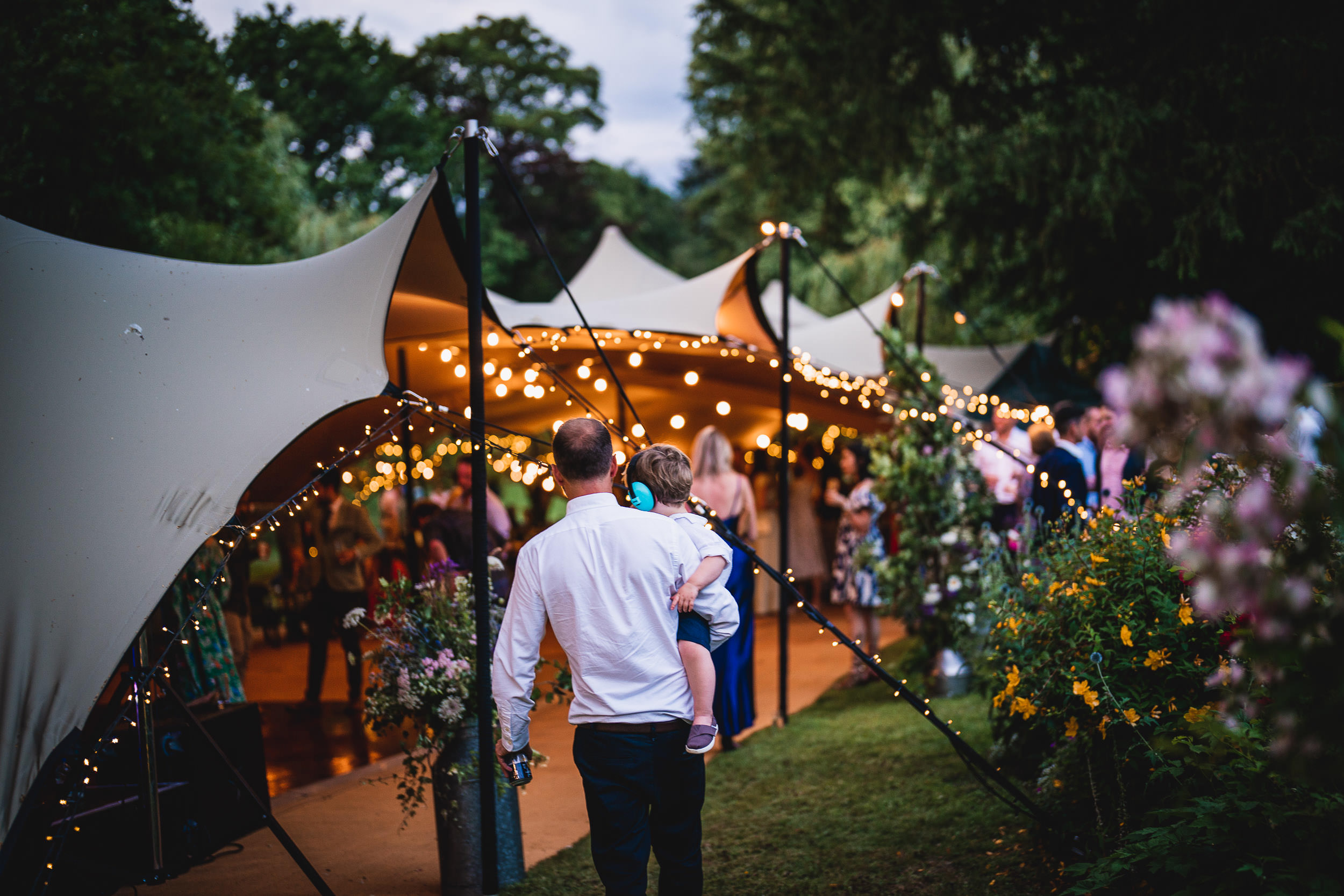 A man carrying a child walks towards a tent decorated with string lights at an outdoor event, with people mingling nearby.