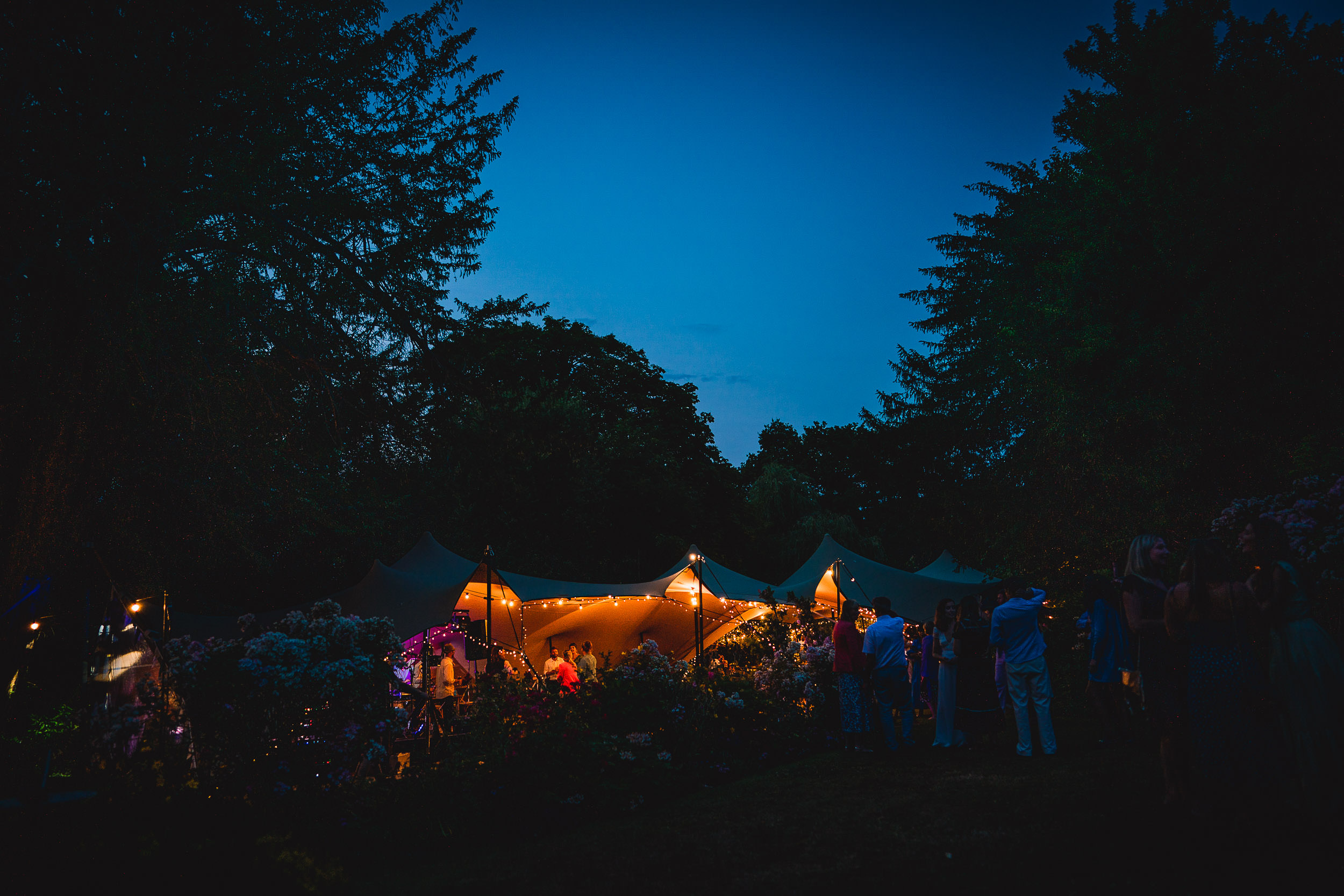 A nighttime outdoor gathering with a tent lit by warm lights. People are gathered around, with a dark blue sky and trees surrounding the area.