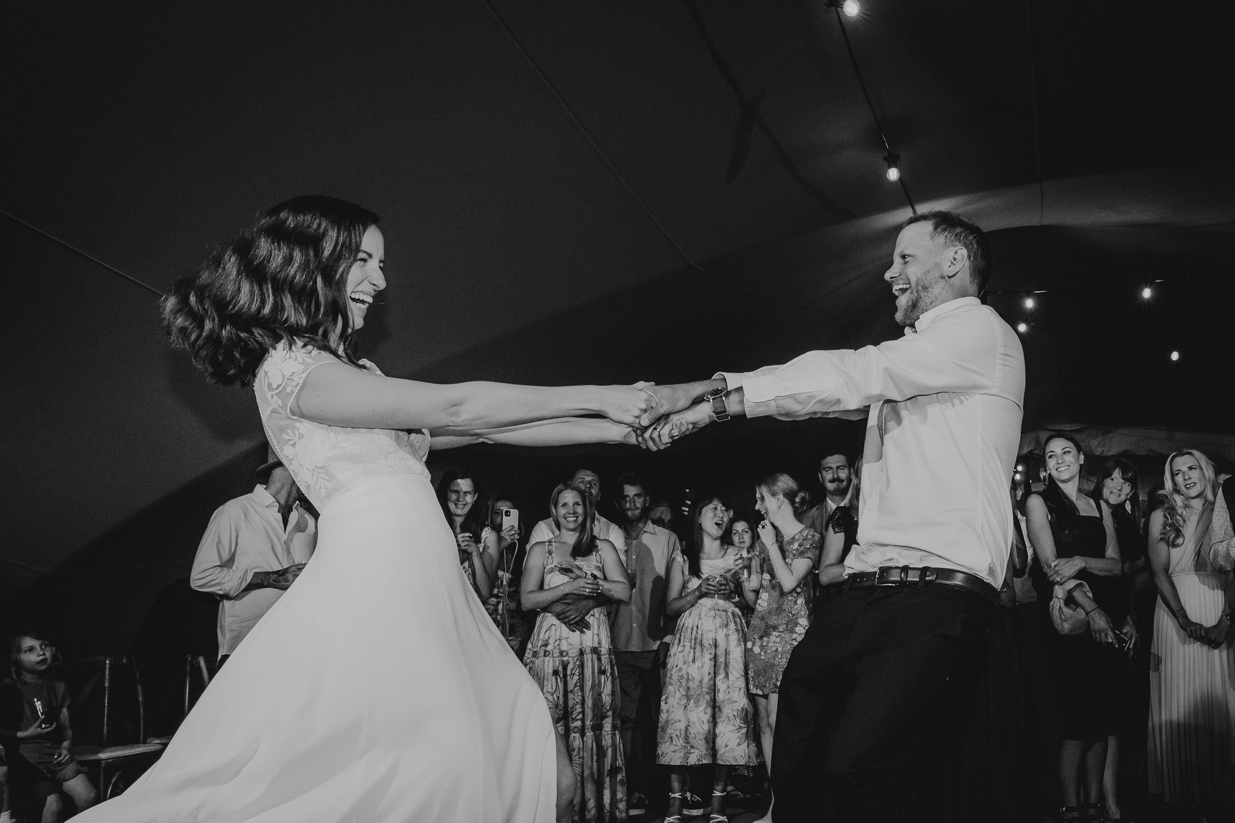 A bride and groom dance joyfully at their wedding, surrounded by smiling guests under string lights.