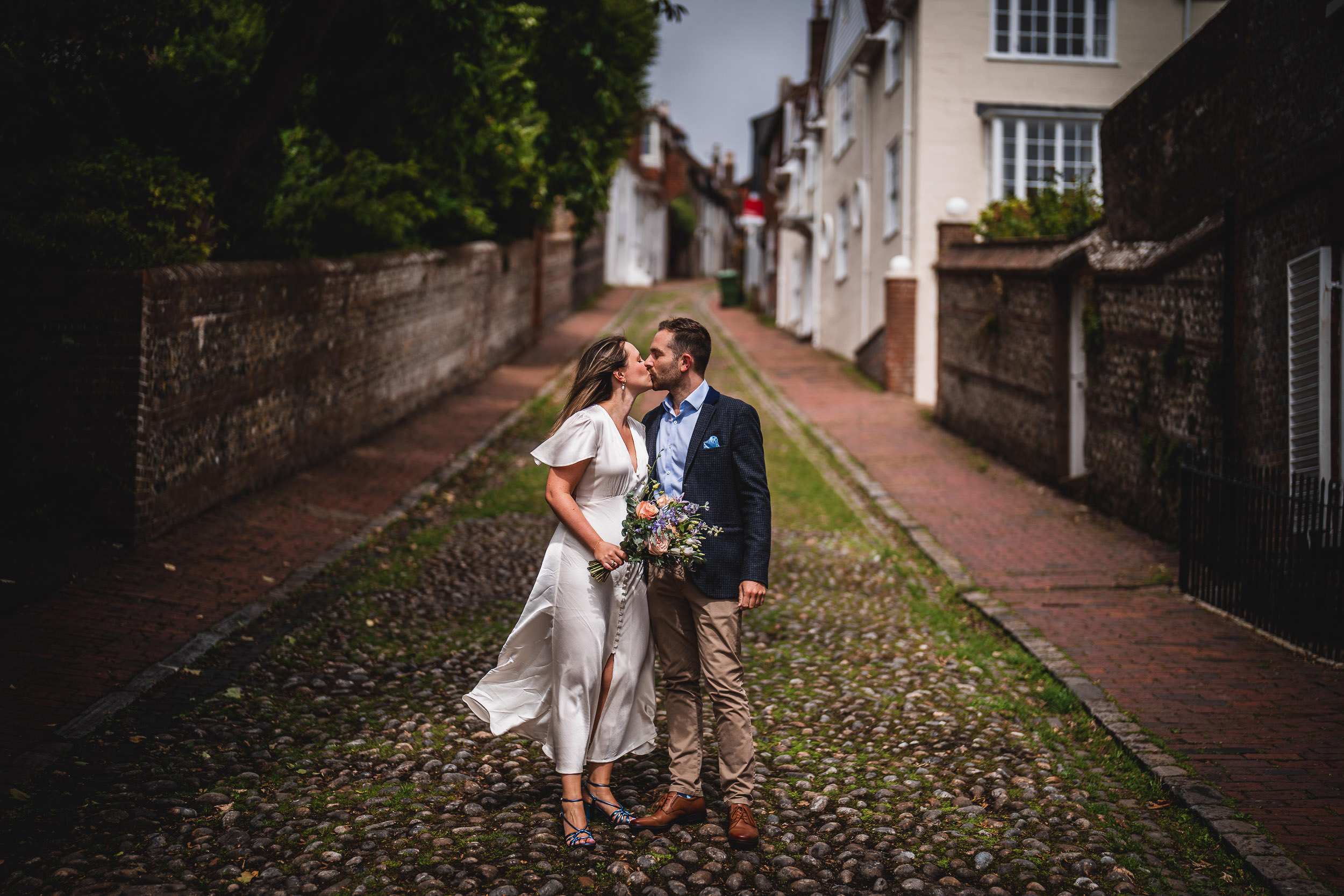 A couple kisses on a cobblestone street, with the woman in a white dress holding a bouquet and the man in a suit. Houses and trees line the background.