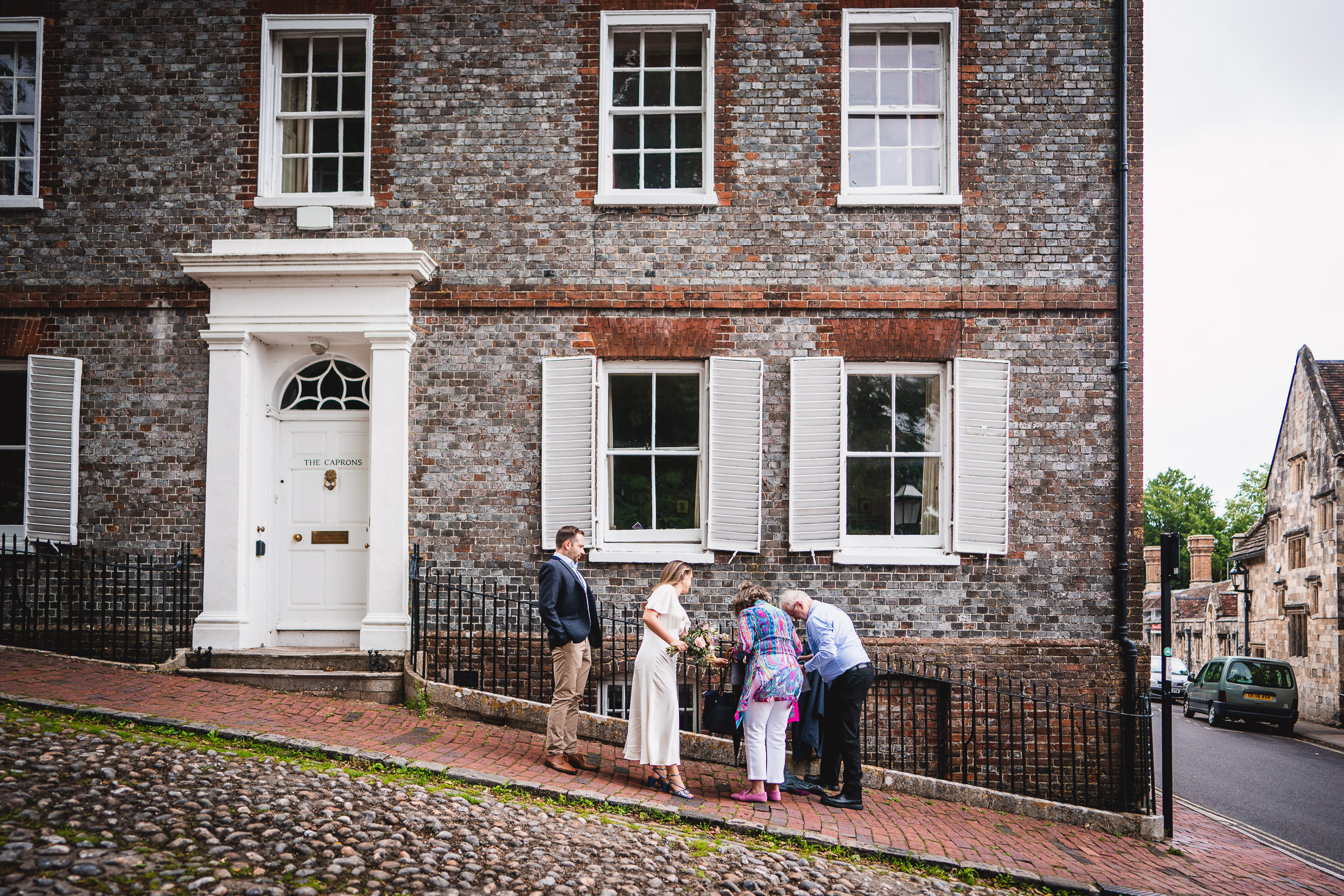 A group of people stand on a cobblestone street outside a brick building with white shutters and a door labeled "The Chantry.