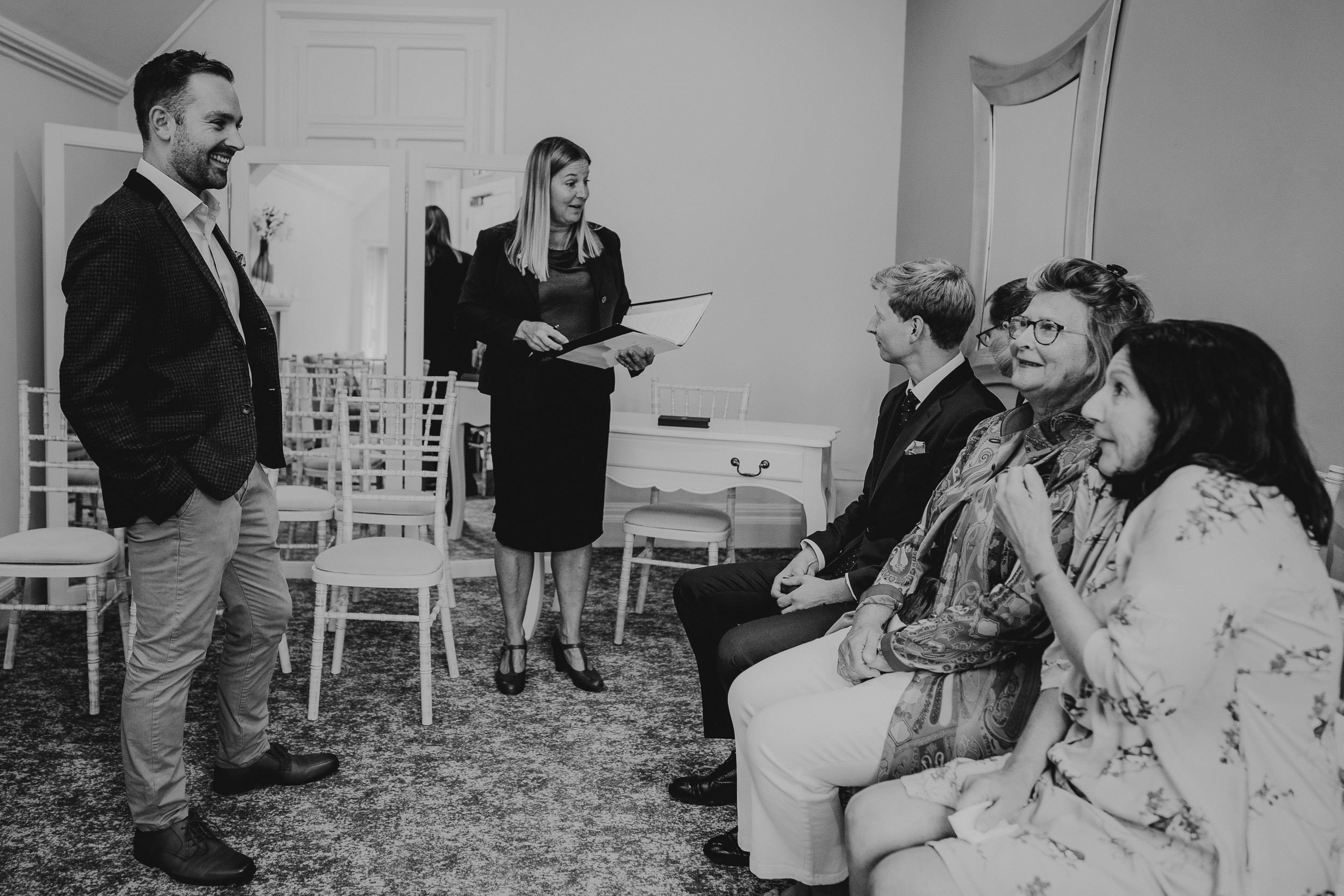 A group of people at a small indoor gathering. One person is standing and speaking, while others are seated and listening. Chairs and a mirror are visible.