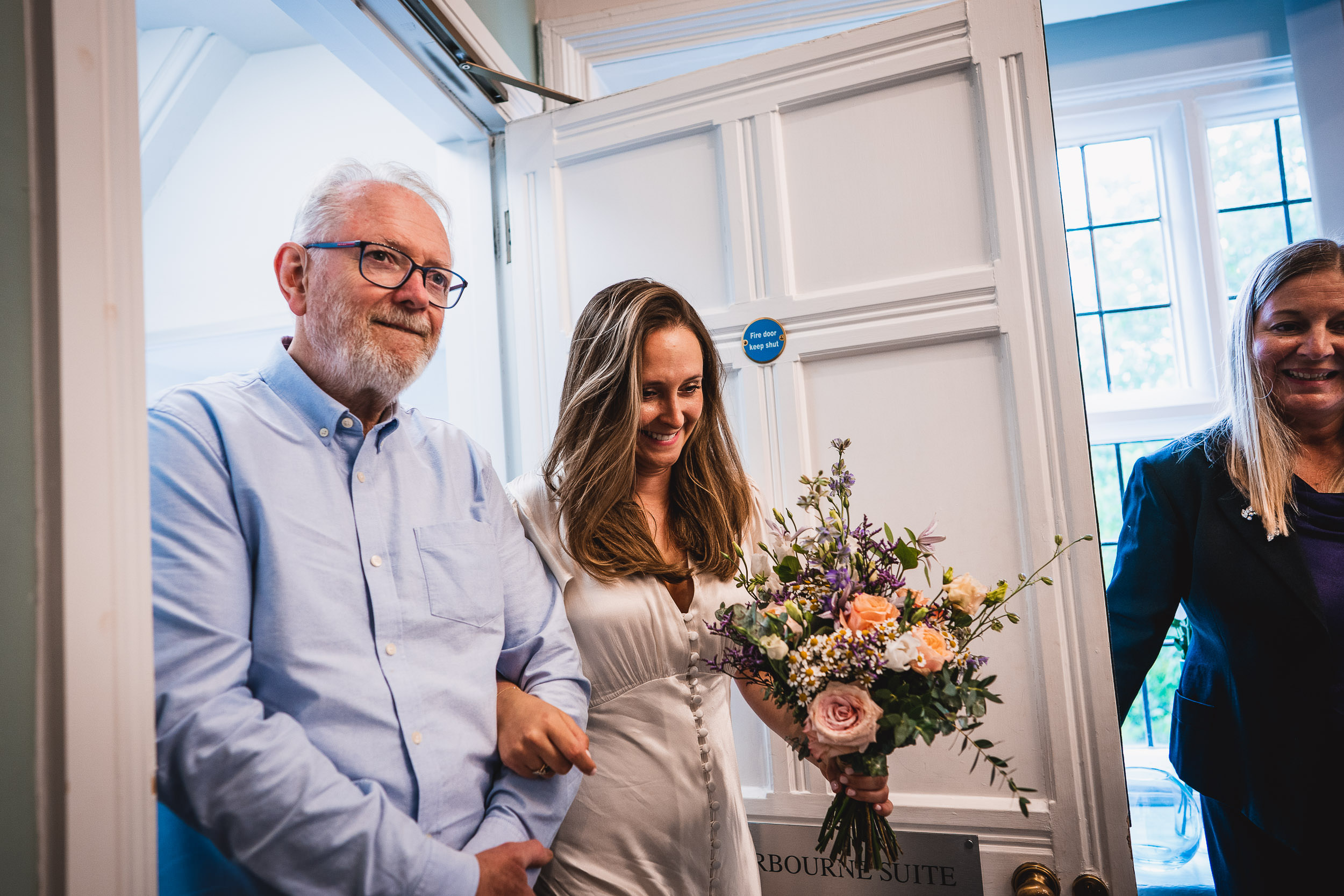 A bride smiles while holding a bouquet and is escorted by a man in a blue shirt through a doorway. Another woman stands nearby.