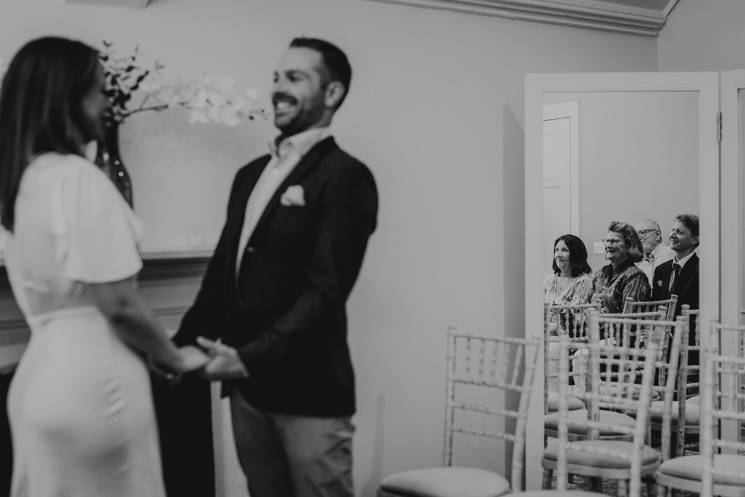 A couple holds hands smiling during a ceremony with four seated people visible in a mirror reflection.