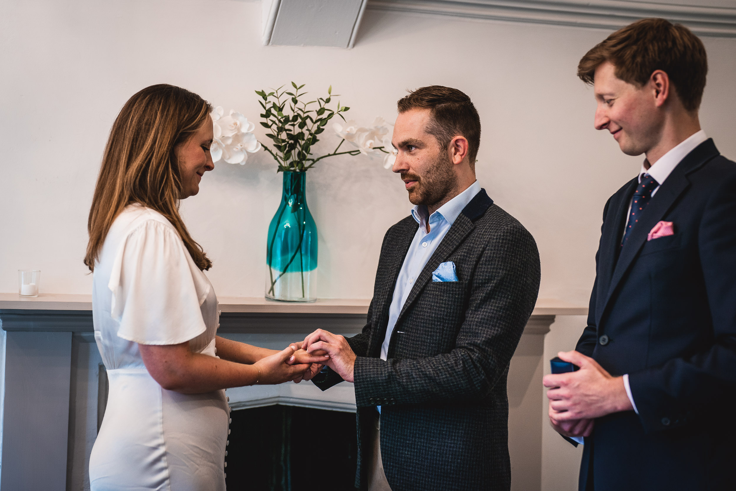 A couple exchanges rings during a wedding ceremony with an officiant standing nearby. A vase with flowers is in the background.