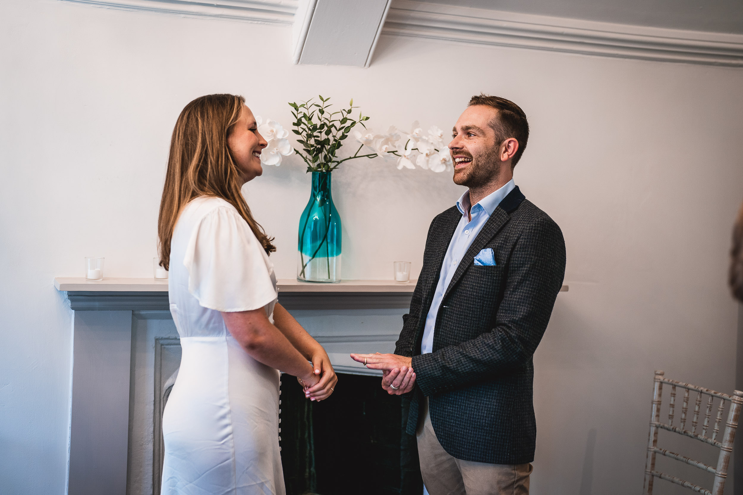 A couple stands facing each other in a white room, smiling and holding hands. A blue vase with white flowers is on a mantel behind them.