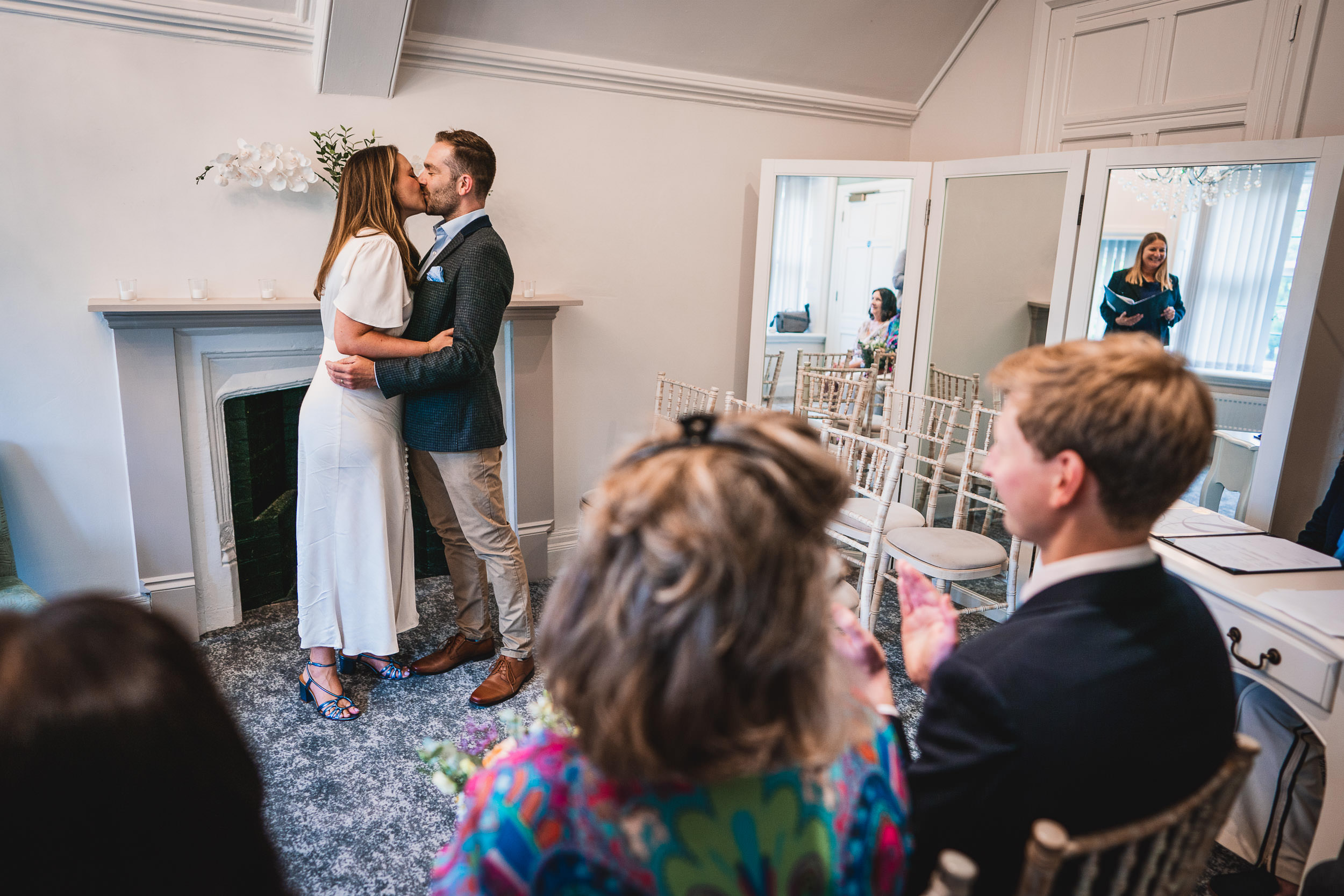 A couple kisses at their wedding ceremony in a small, decorated room, with guests seated and clapping.