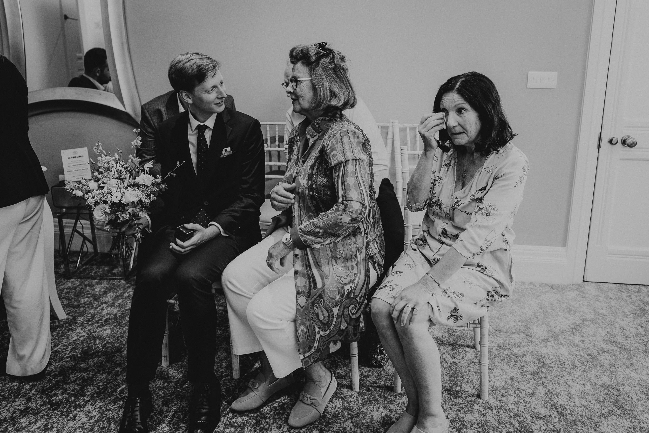 A man in a suit sits with two women on chairs in a room. One woman holds a bouquet, and the other wipes her eye.