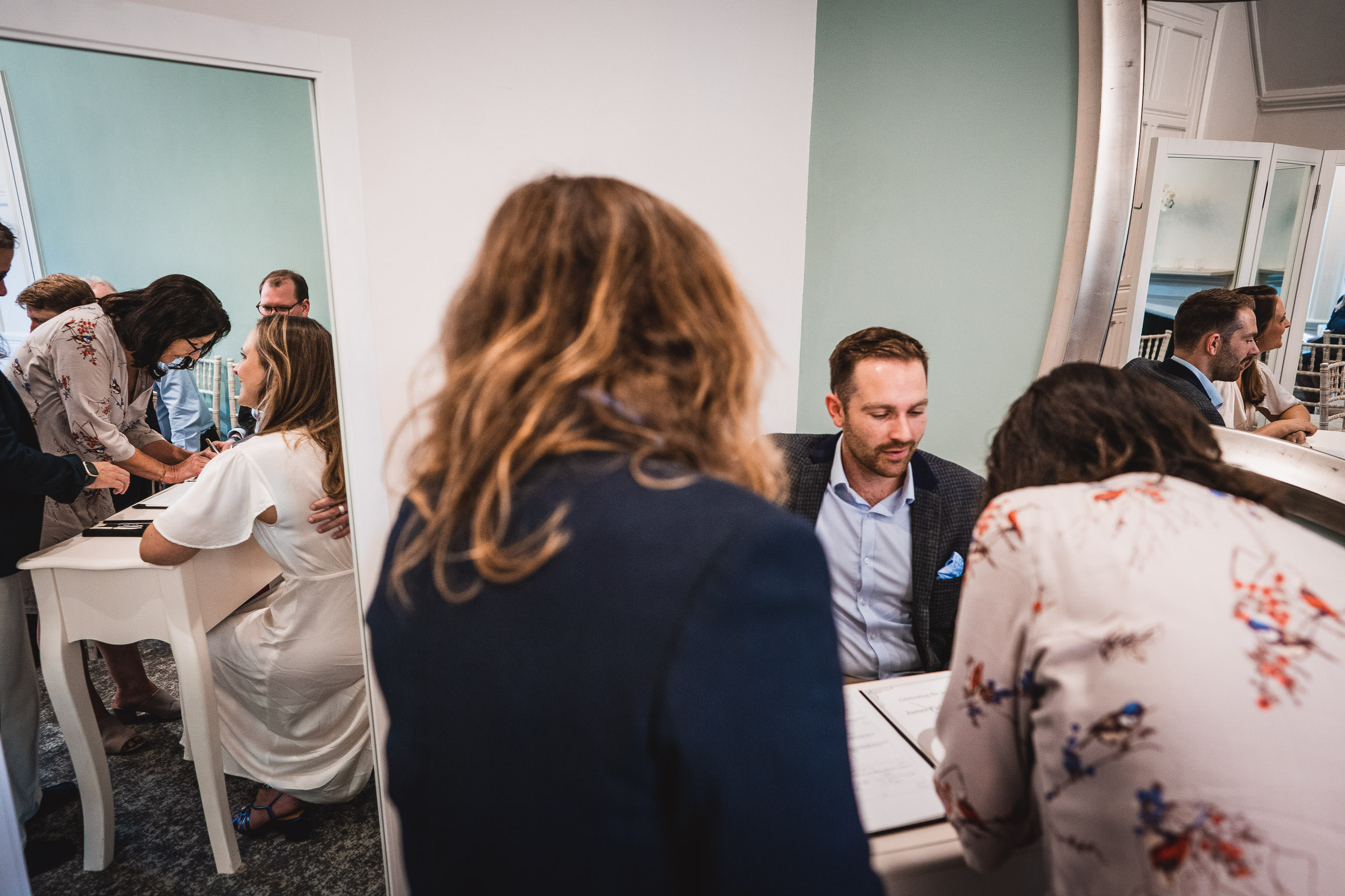 People in a room are gathered around tables, engaging in conversation and writing. A large mirror reflects the scene.