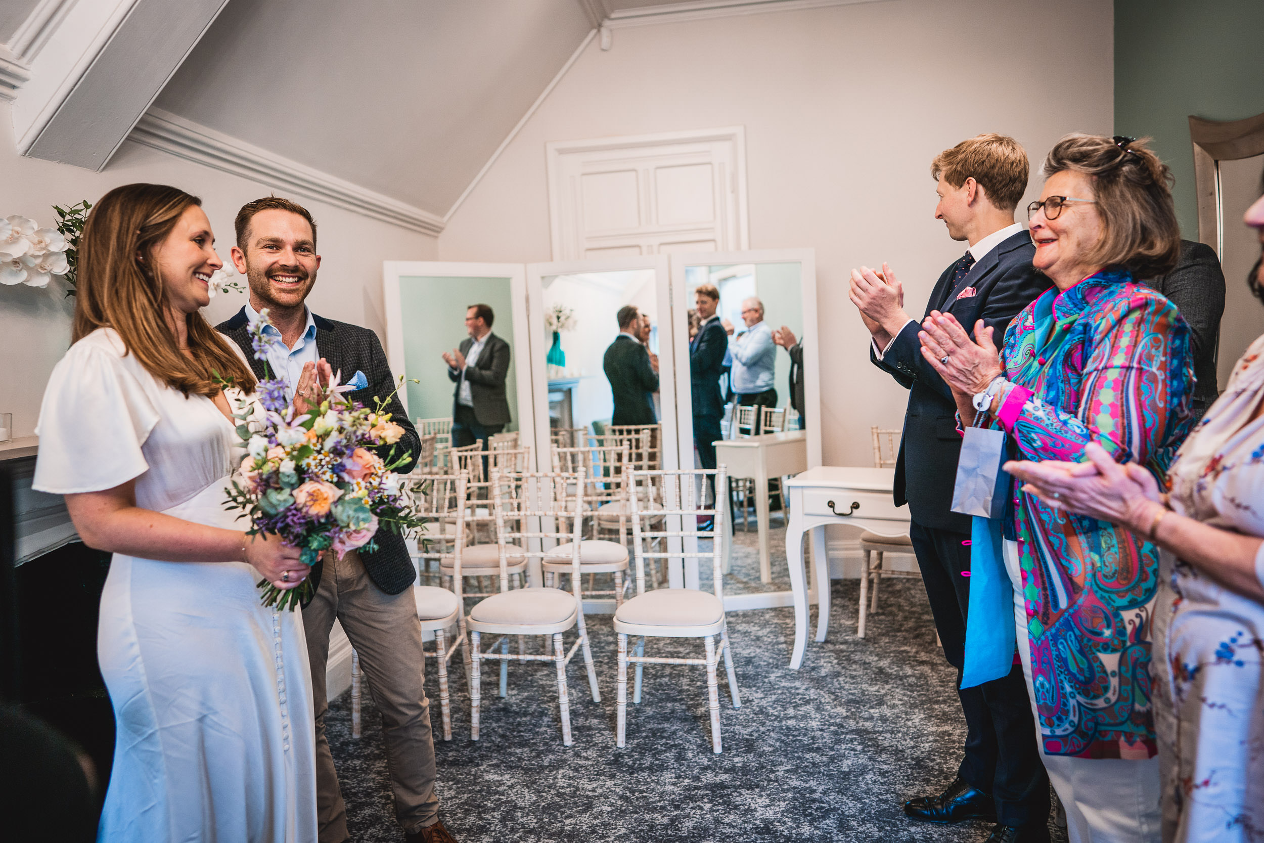 A couple stands facing guests in a small room, holding a bouquet. Guests are clapping and smiling. White chairs are arranged behind the couple. Large mirrors reflect the scene.