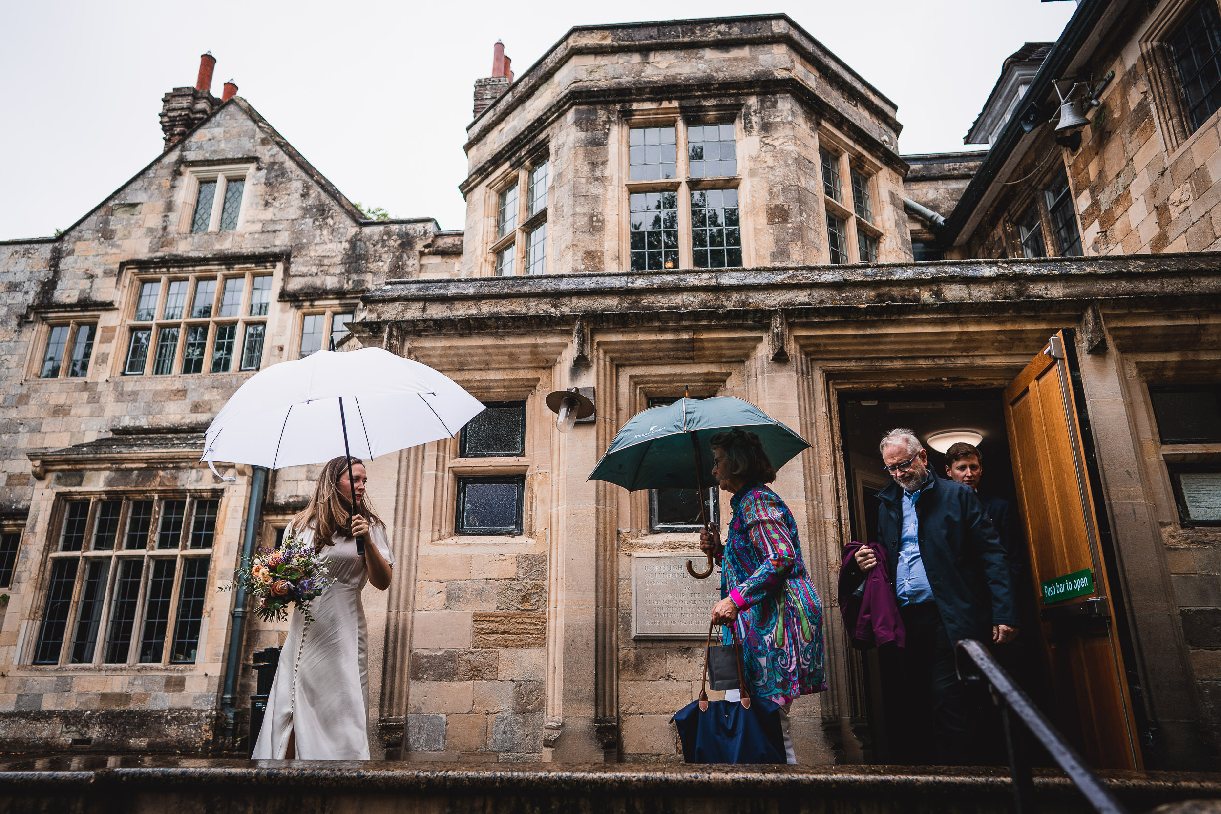 A bride holding a bouquet and a white umbrella stands outside an old stone building. Three people with umbrellas exit the building under a cloudy sky.