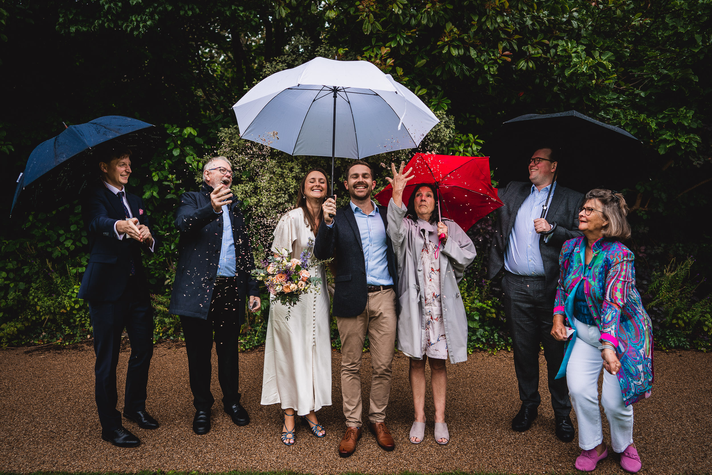 A group of people smiling under umbrellas in the rain, with some throwing confetti. They are on a path surrounded by greenery.