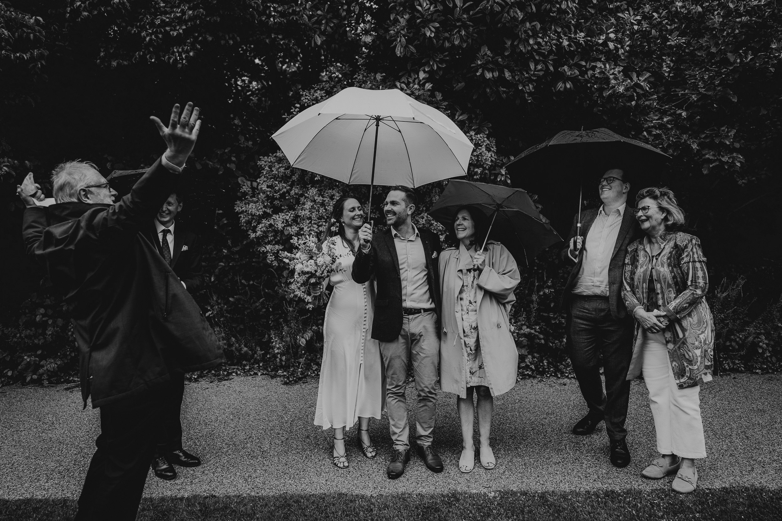 Group of people standing outdoors in the rain, holding umbrellas. A man gestures enthusiastically at them.