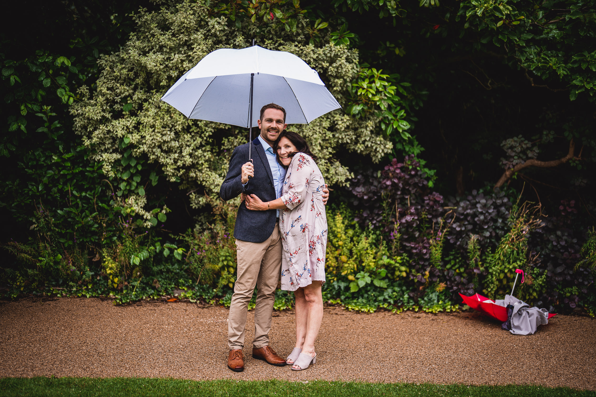 A couple stands under a white umbrella in a garden. The man is wearing a dark blazer and khaki pants, and the woman is in a floral dress. A closed red umbrella is on the ground nearby.