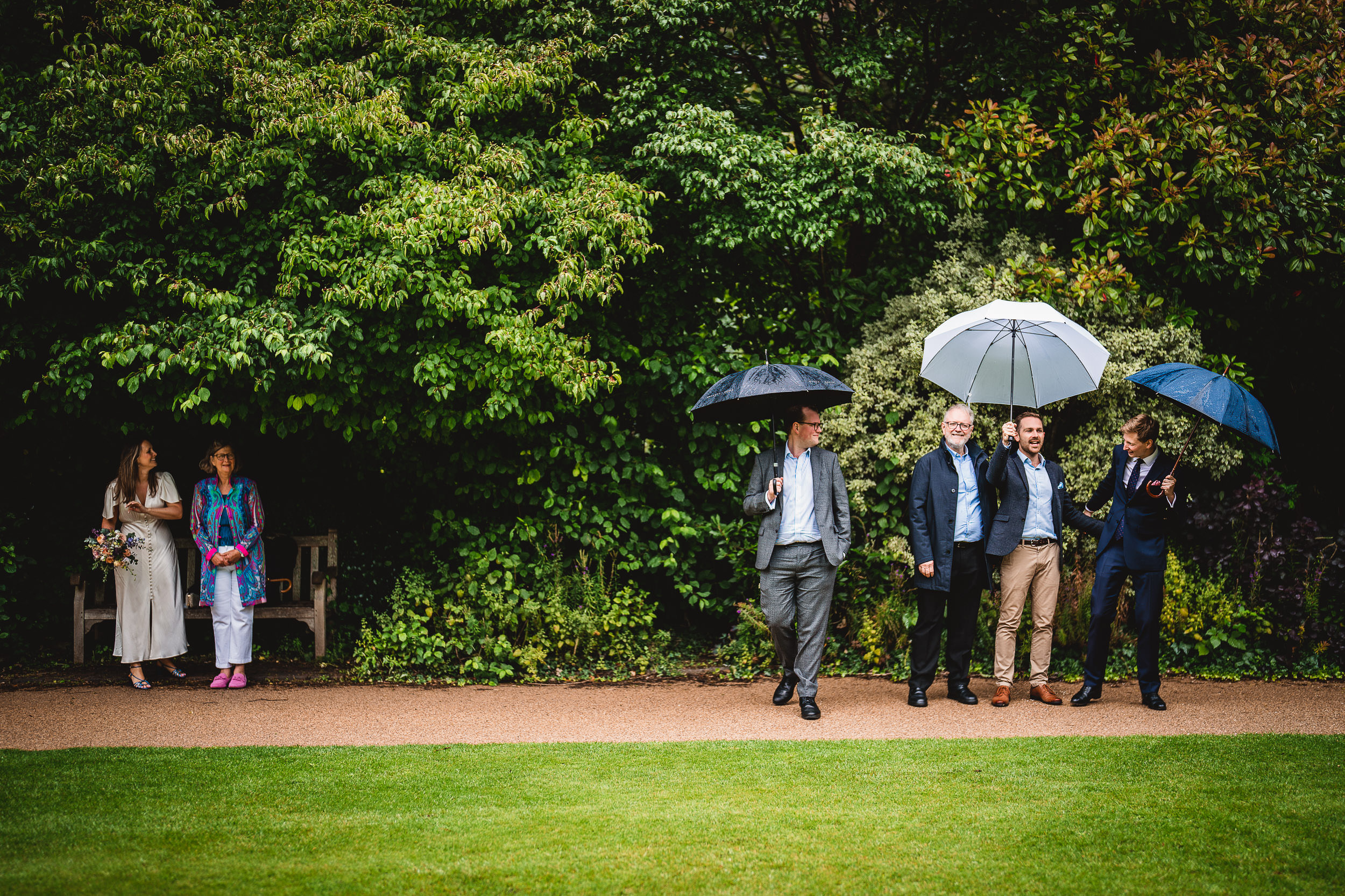 A group of people stand on a path in a garden, under umbrellas. Two are sitting on a bench to the left, and four are standing on the right. It appears to be a rainy day.