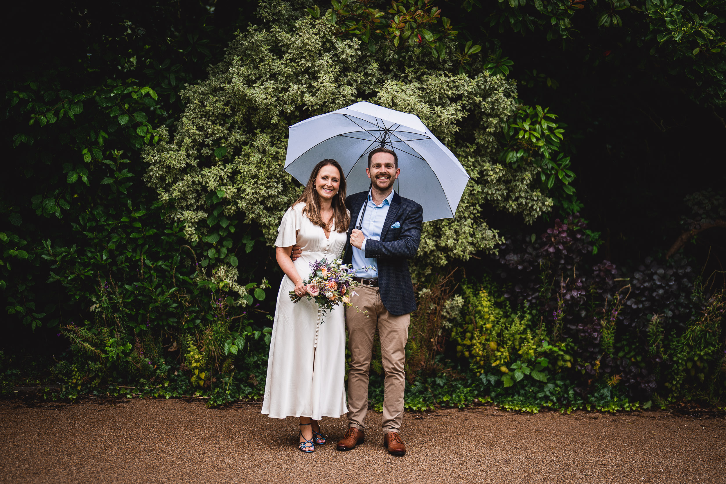 A couple stands under a white umbrella in a garden, with the woman holding a bouquet.