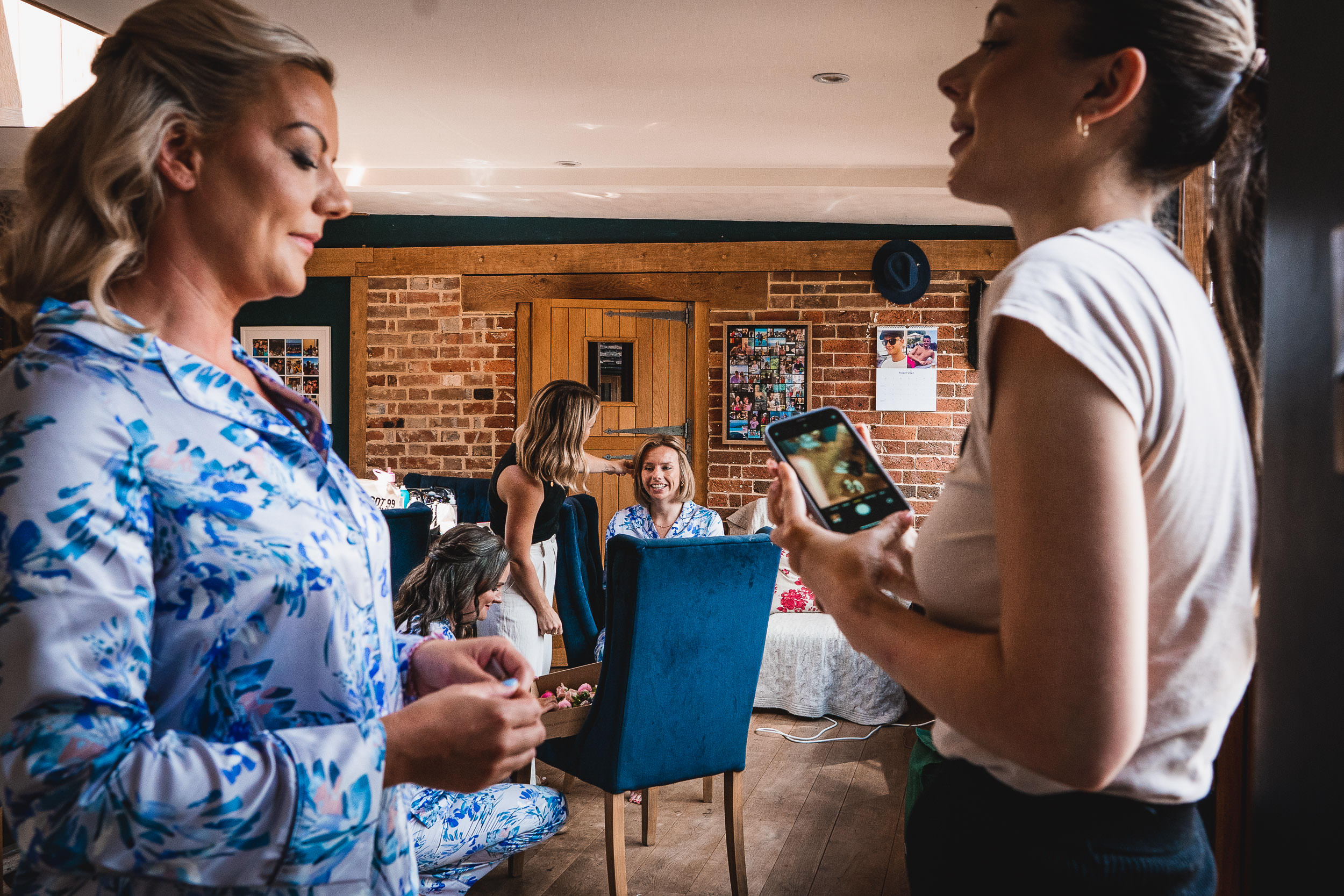 People interact in a room with brick walls and wooden flooring. Some are talking, one is holding a phone, and others are seated. There are photos and a hat on the walls.