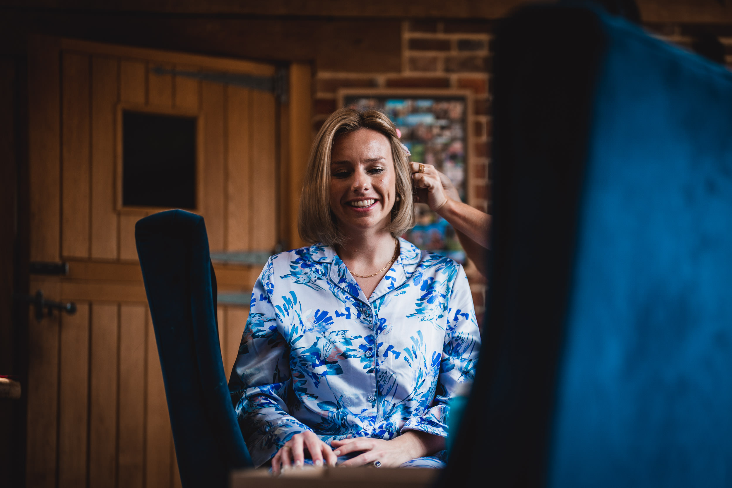 A woman in floral pajamas sits indoors with a person styling her hair.