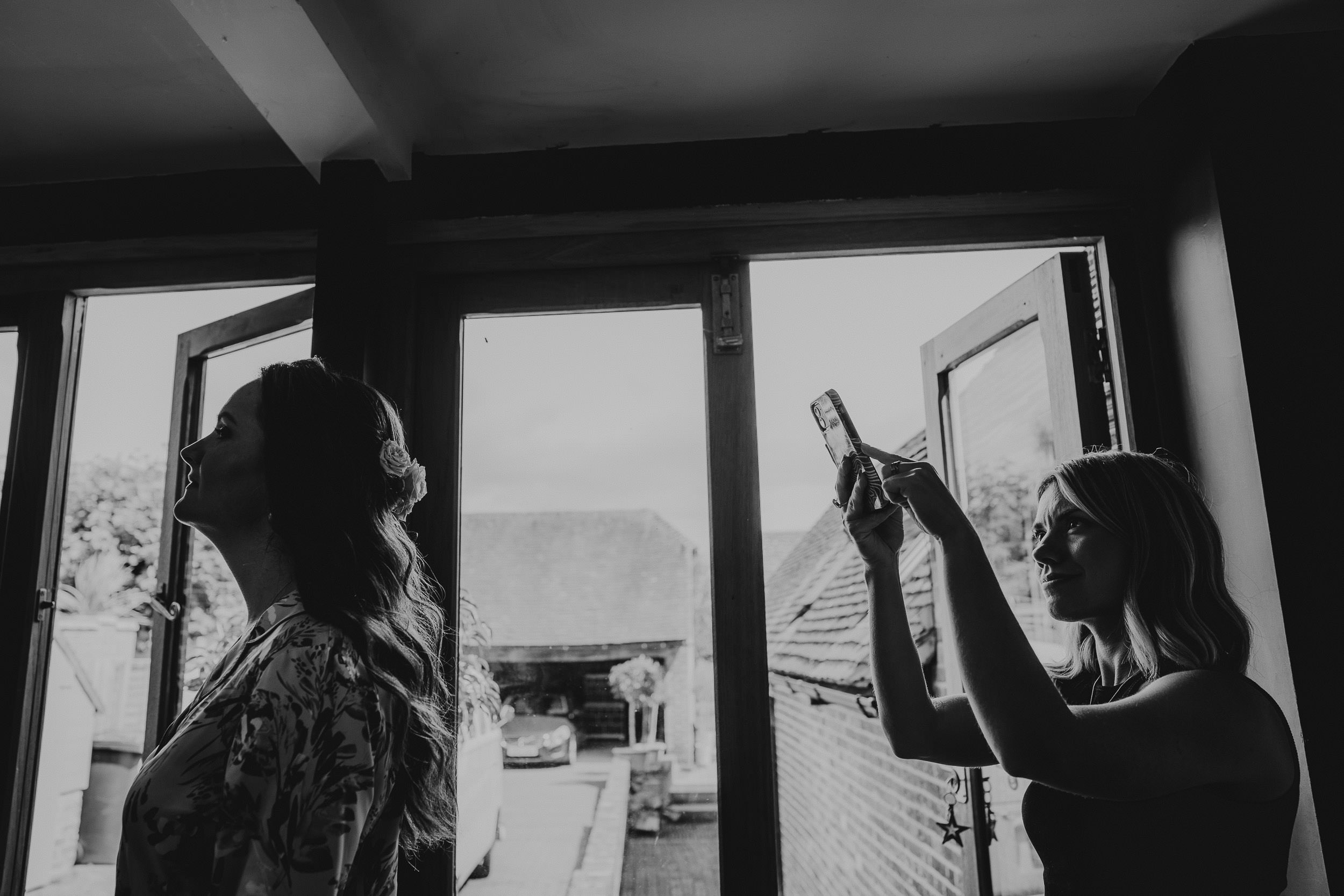 Two women indoors near open windows. One woman is taking a photo with her phone while the other stands facing away, smiling. The background shows a driveway and buildings outside.