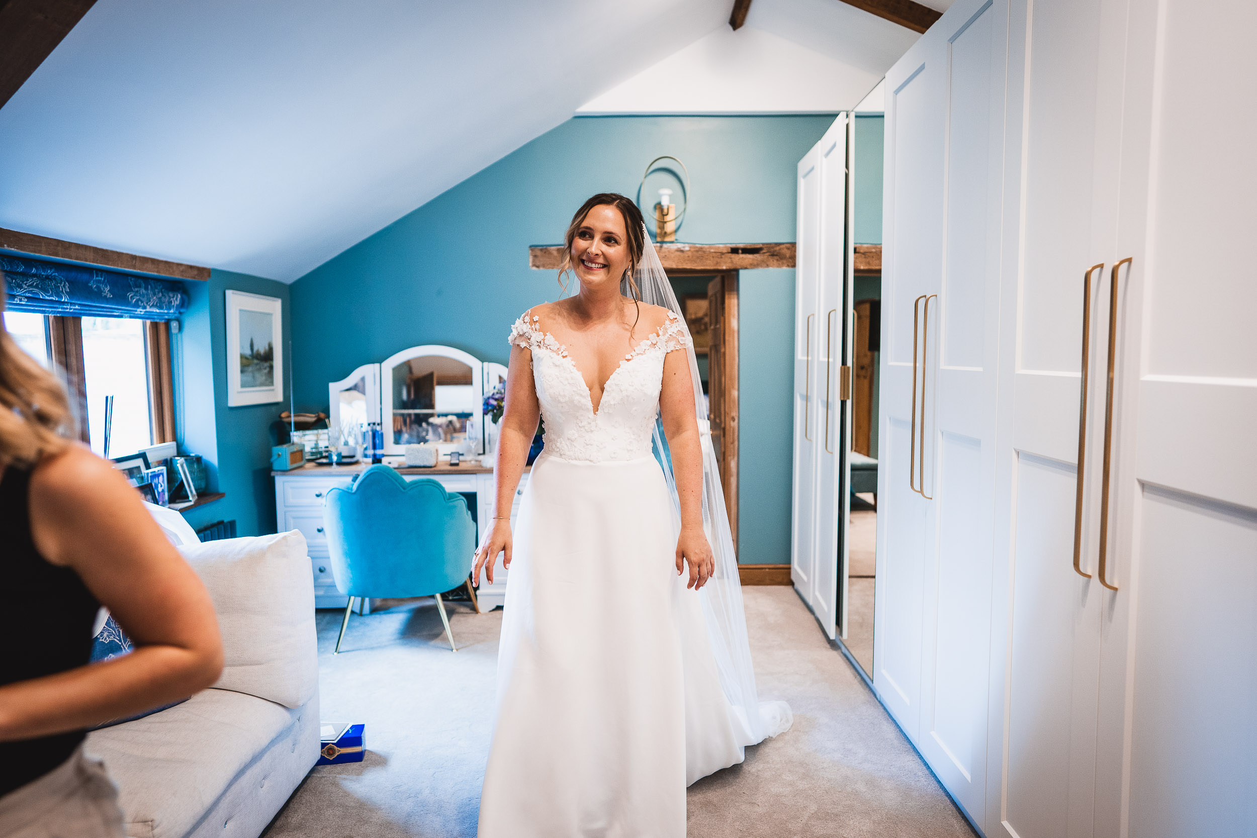 A bride in a white dress smiles while standing in a bright room with blue walls and a dressing table.