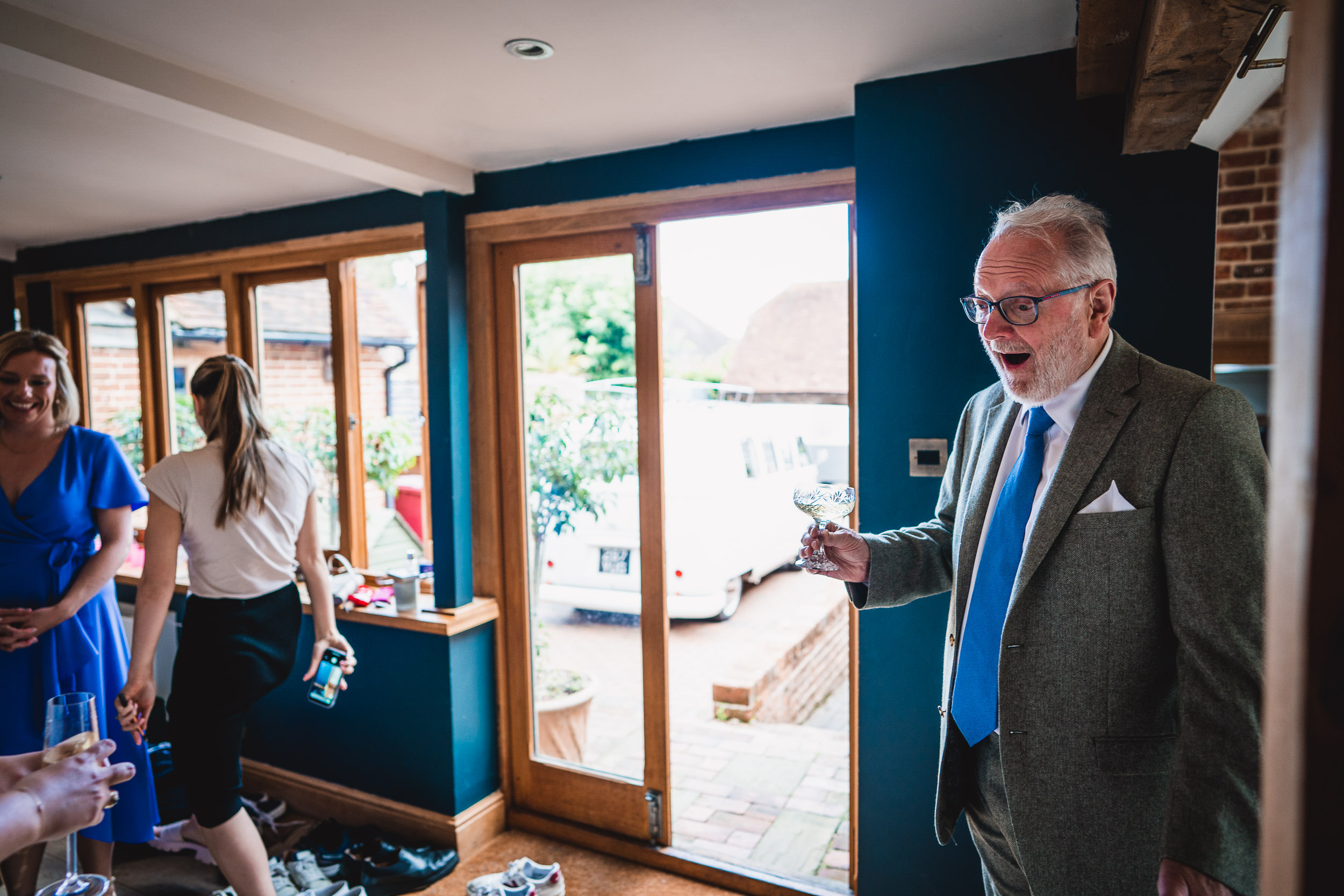 An older man in a suit holds a glass, looking surprised. Two women stand near a window with shoes on the floor.